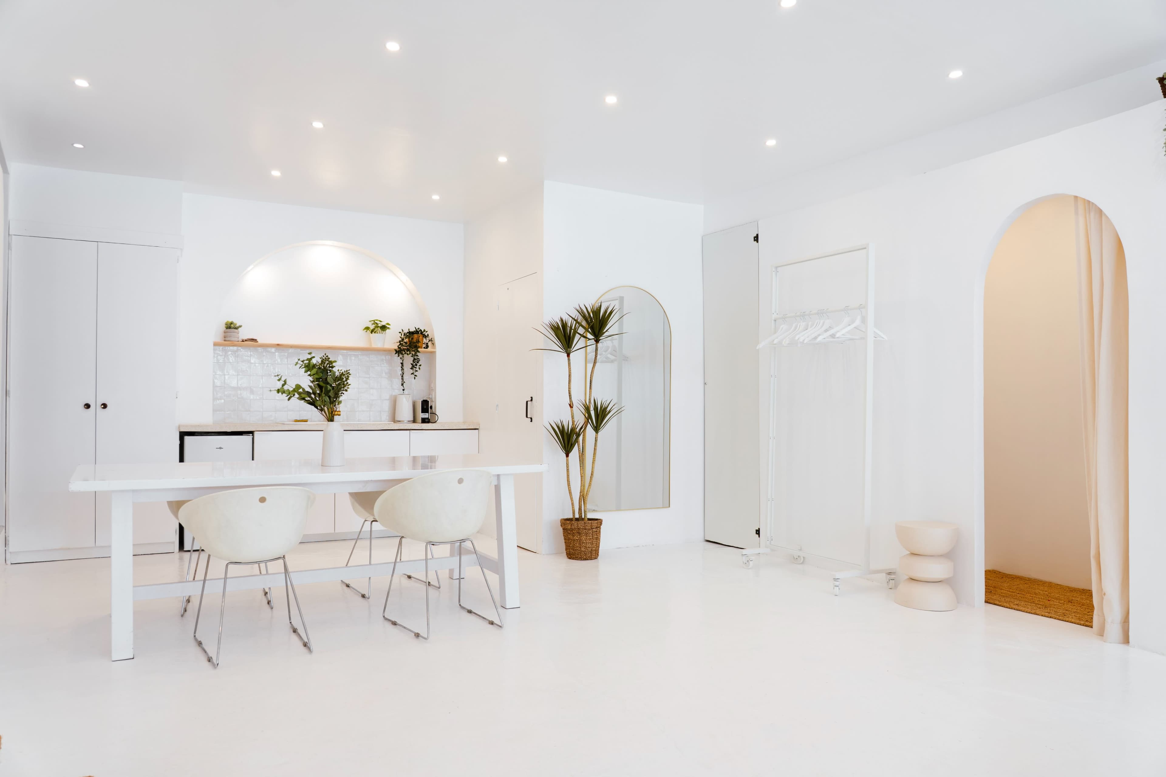 The image shows a modern, minimalist interior with a white dining table, two chairs, a small kitchenette area, and light-colored decor.