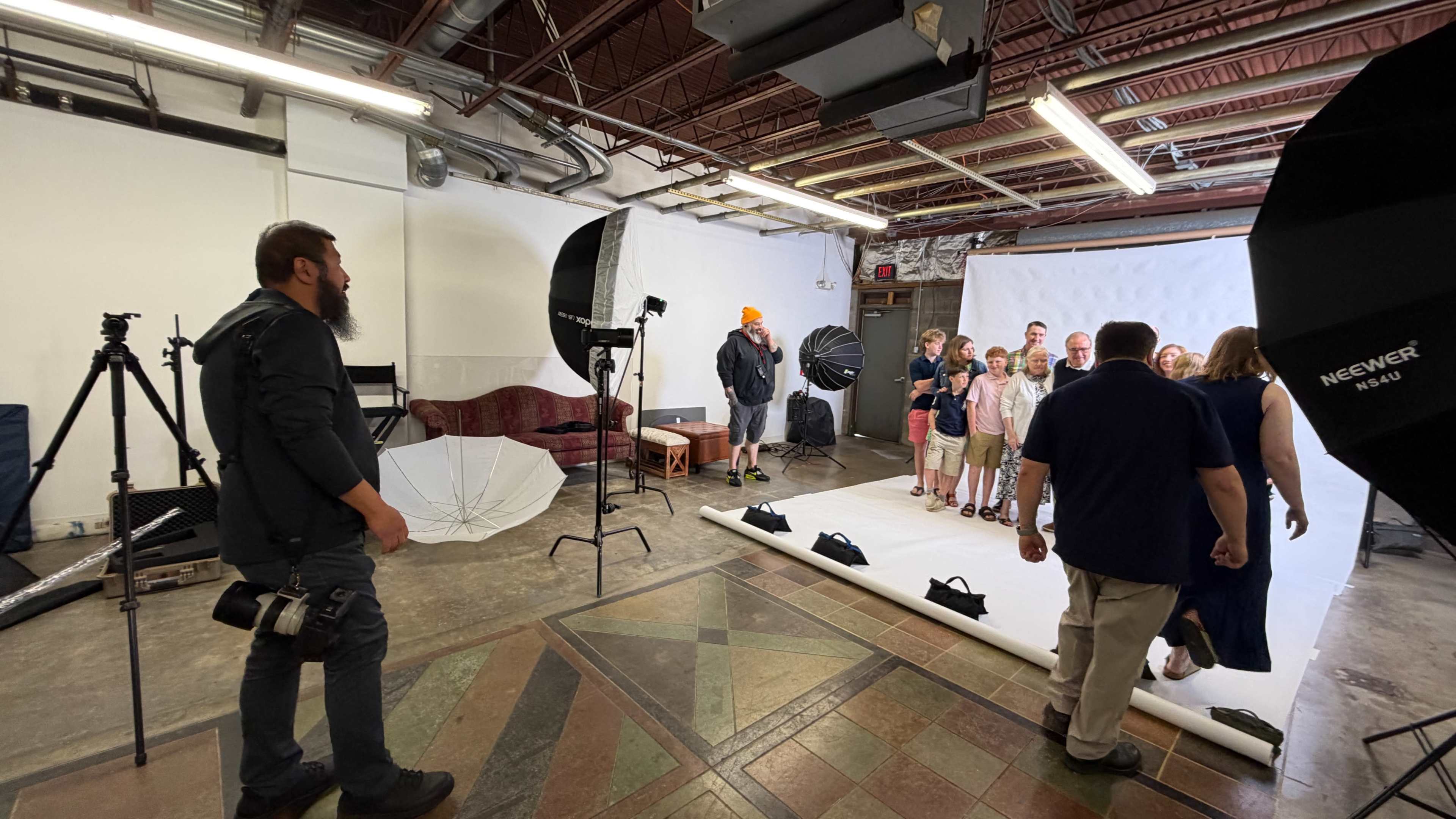 A photographer sets up equipment in a studio while a group of people prepares for a photoshoot against a white backdrop.