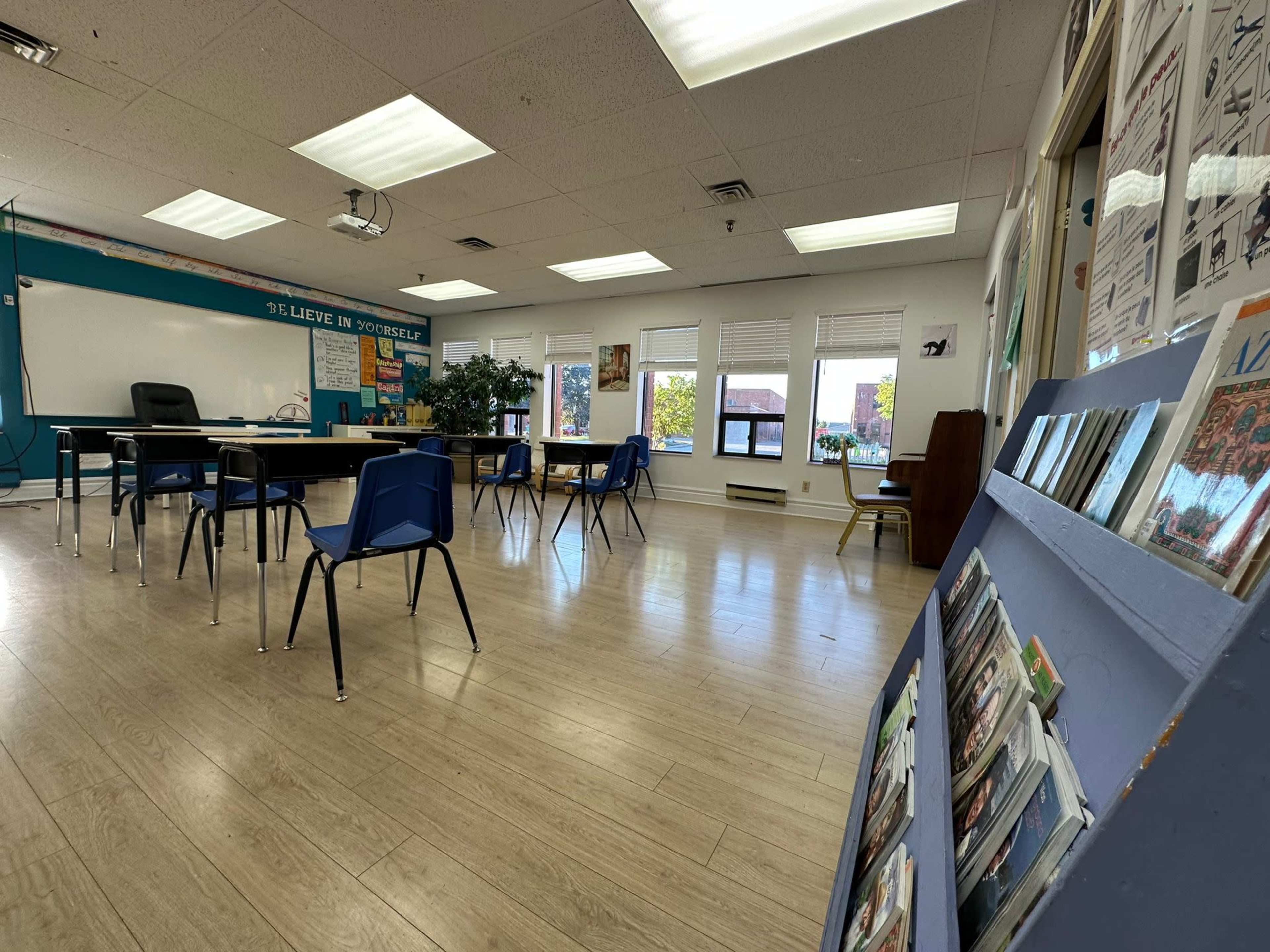 A spacious classroom with blue chairs arranged around a central table and a bulletin board displaying various publications in the foreground.