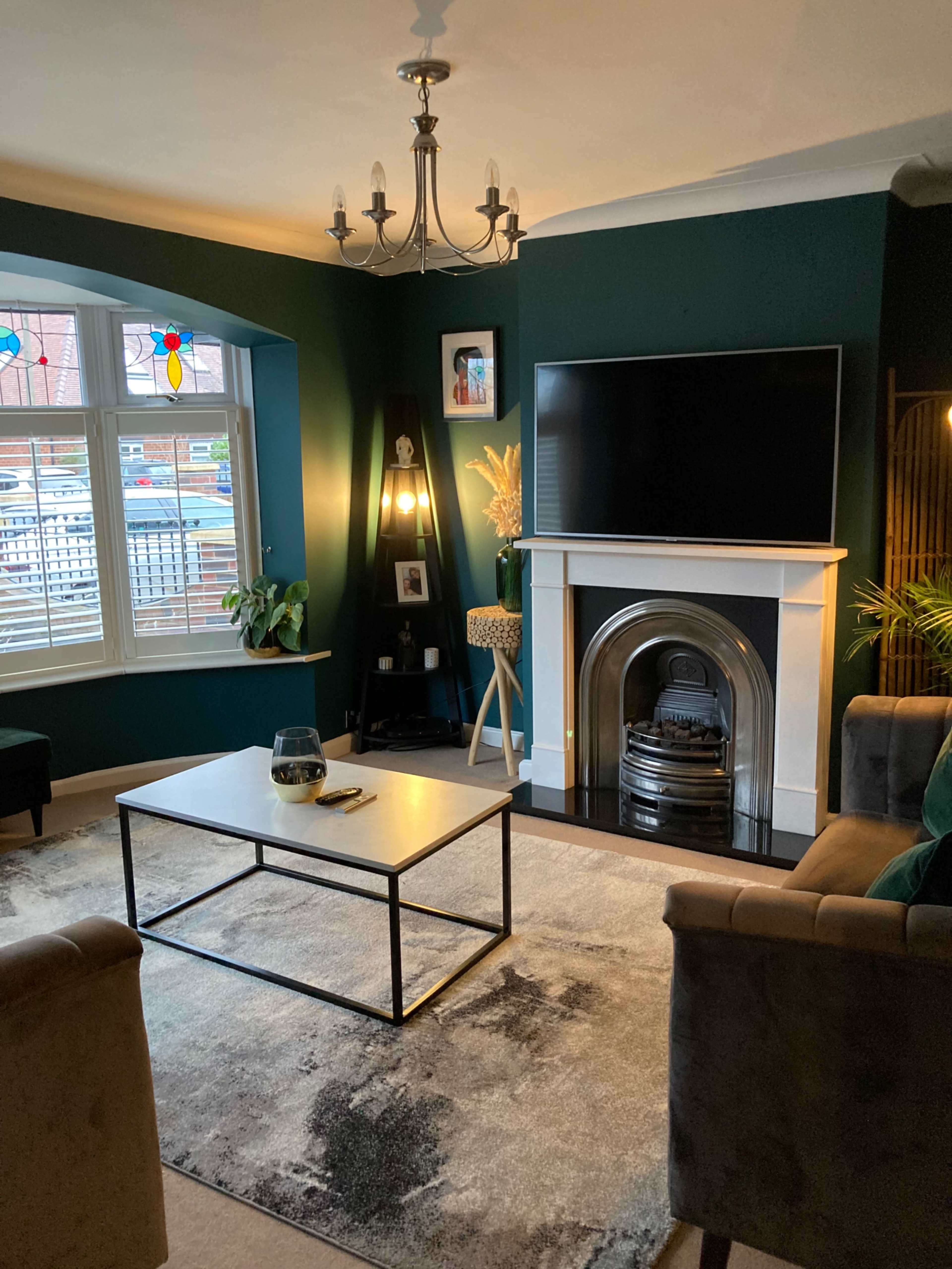 A living room with a dark green wall, a fireplace, a television mounted above the mantle, a coffee table in the center, and two armchairs.