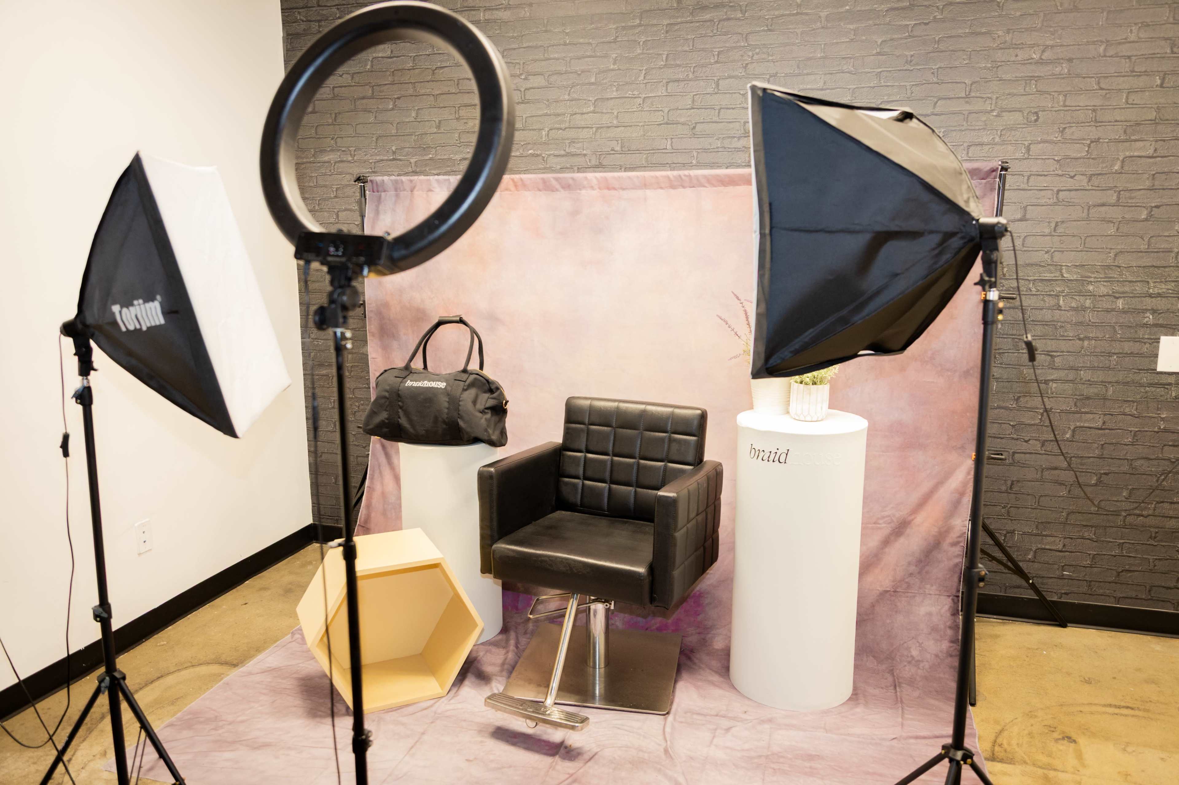 The image shows a photo studio setup featuring a black styling chair, light softboxes, a ring light, and various props including a hexagonal table and a decorative plant on a white pedestal.