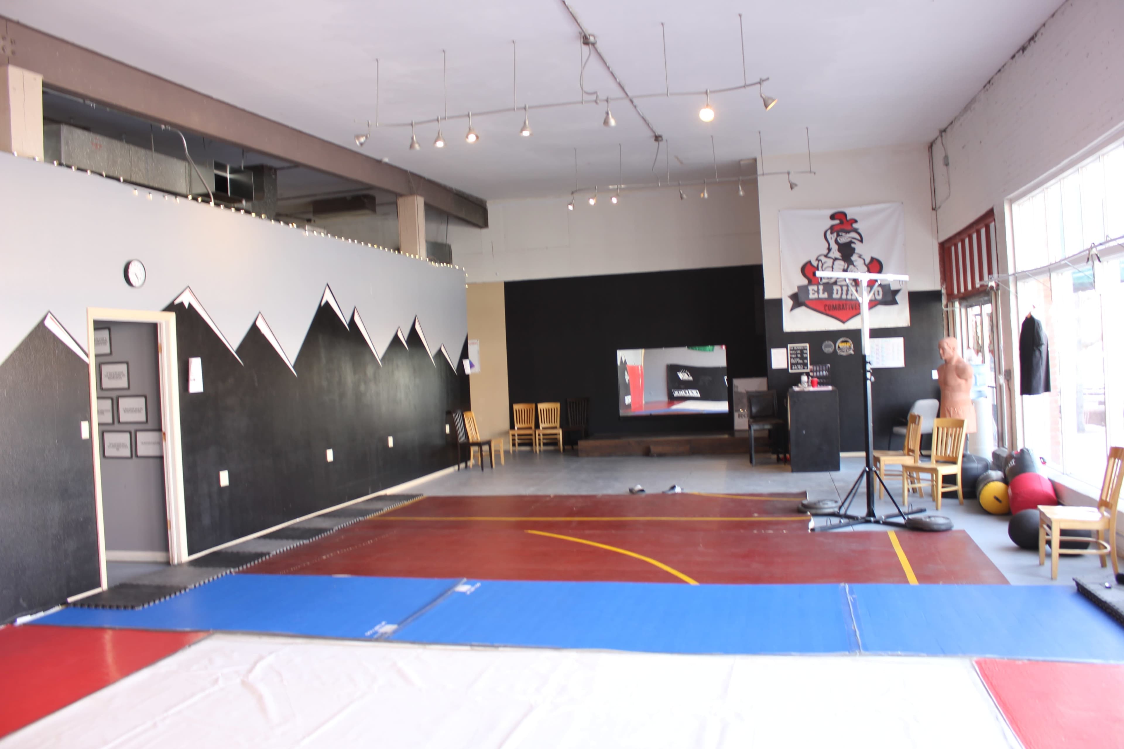 The image shows a spacious martial arts studio with a mix of wooden and padded flooring, black walls featuring a mountain design, and exercise equipment arranged near a large window.
