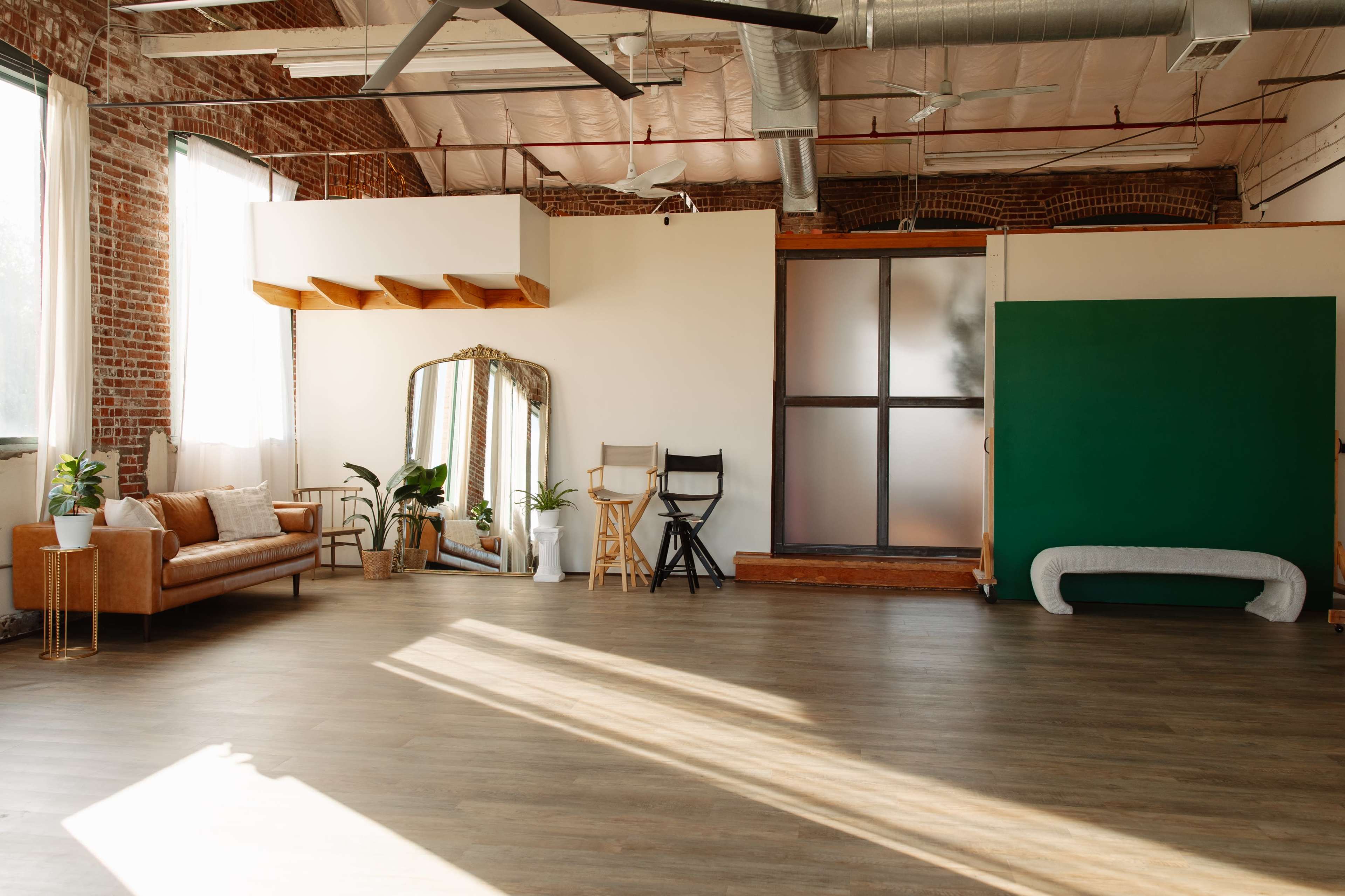 The image shows a spacious, well-lit room with wooden flooring, featuring a brown sofa, a full-length mirror, and a green wall, along with various plants and seating arrangements.