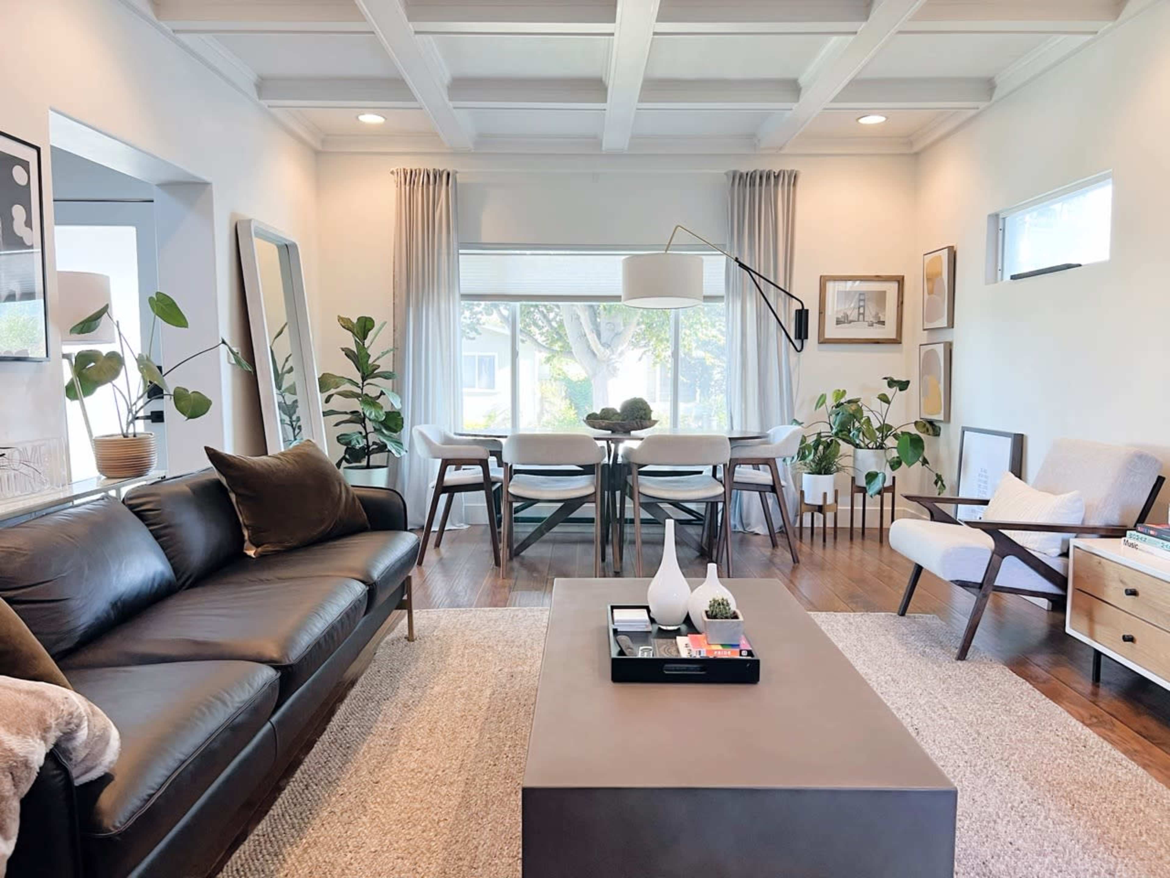A modern living room features a black leather sofa, a coffee table, and a dining area with a table and chairs, surrounded by plants and framed artwork.