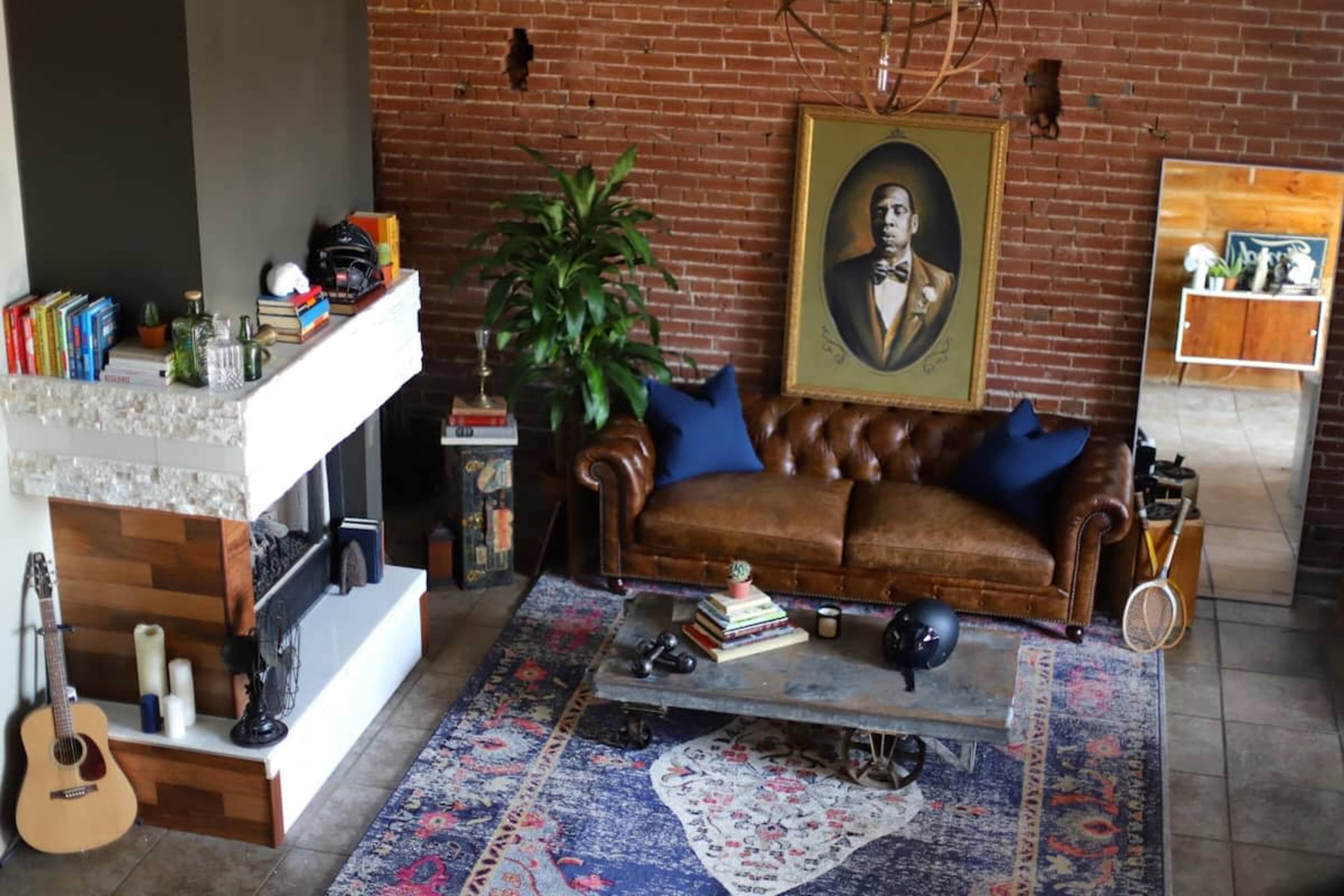 A cozy living room with a brown leather sofa, a large portrait on the wall, a coffee table with books, and a green plant, all set against a brick wall.