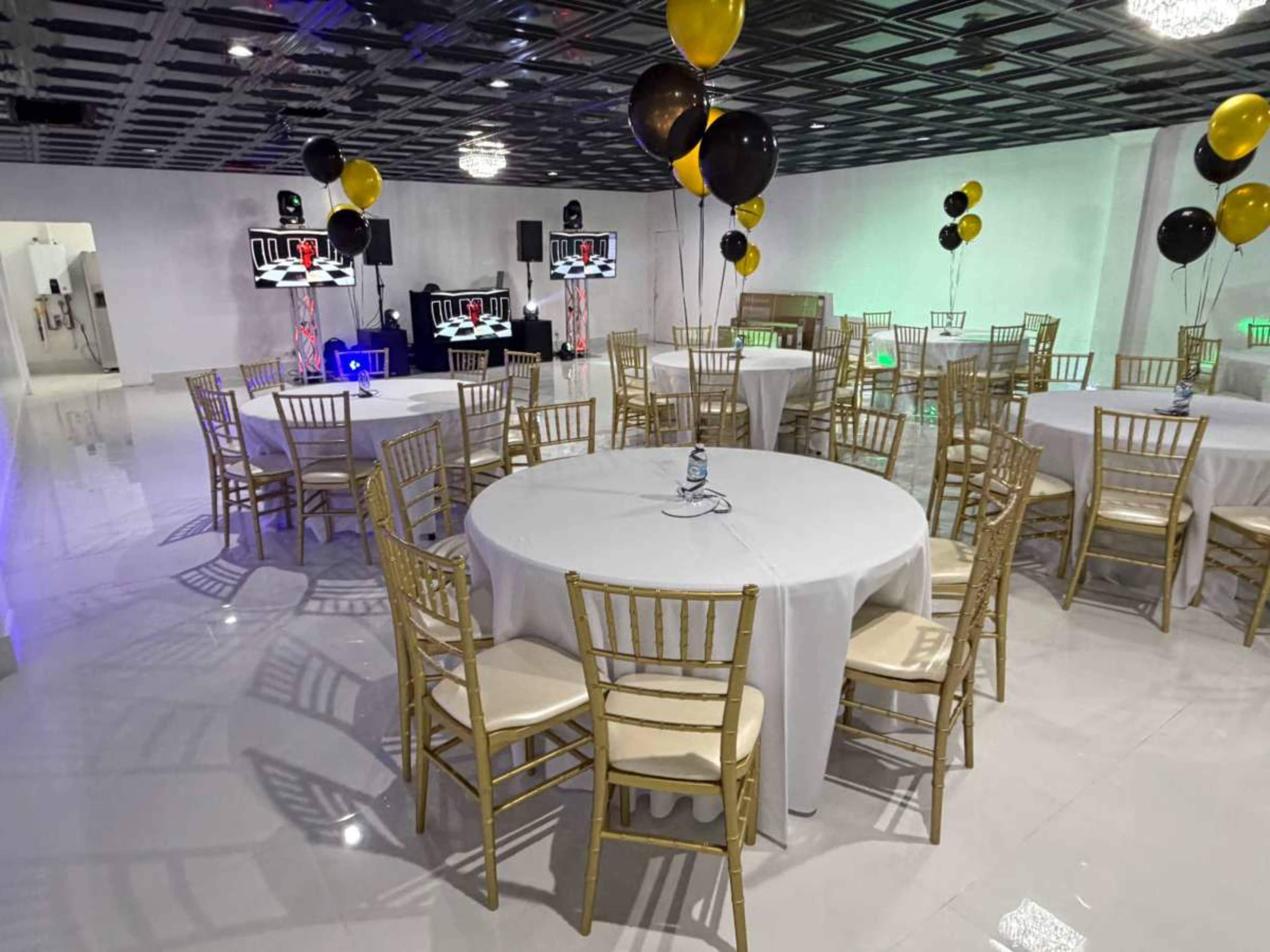 The scene shows a banquet hall set up with round tables covered in white tablecloths, gold chairs around each table, and black and yellow balloons suspended from the ceiling.