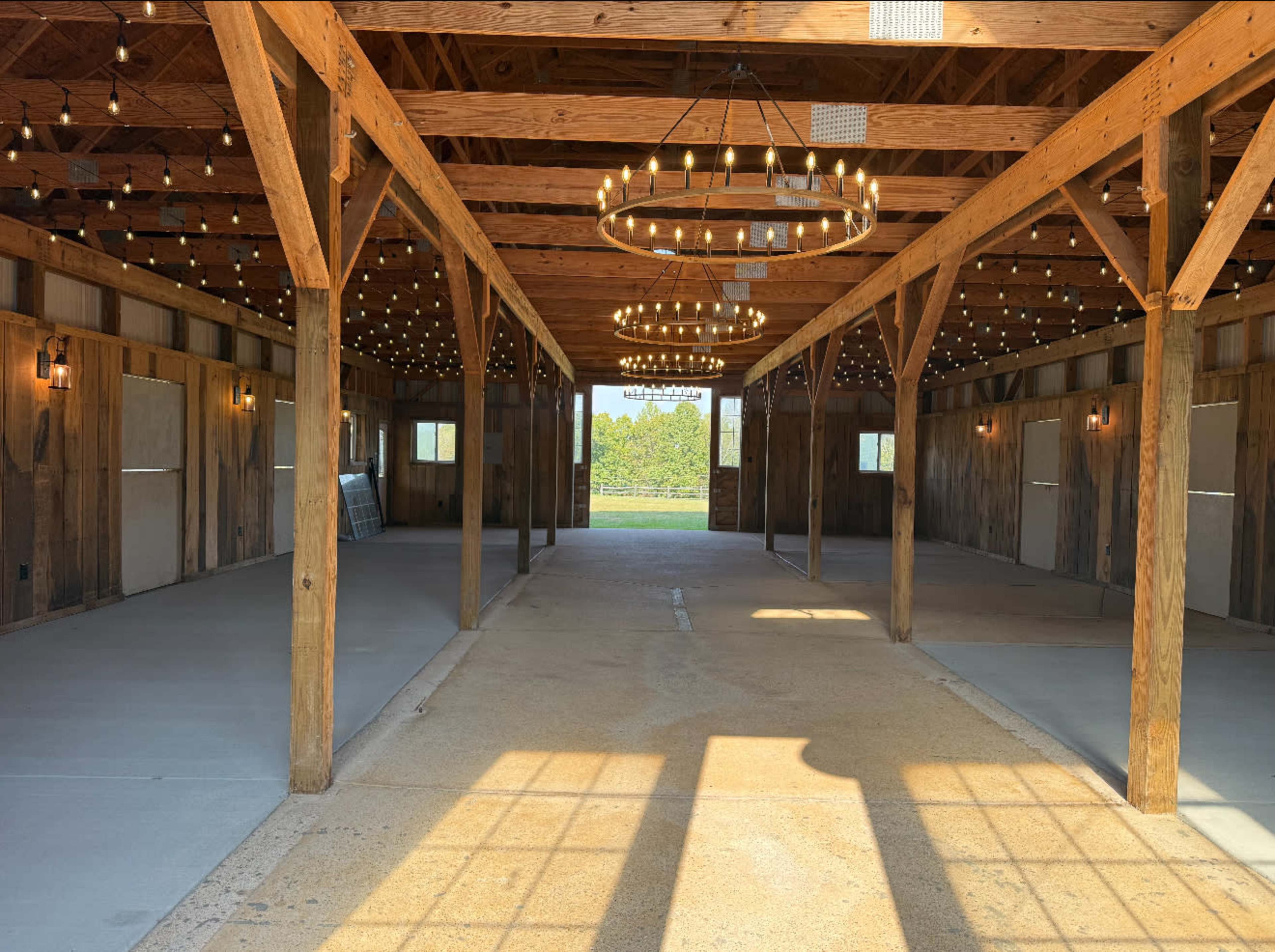 The image shows a spacious barn interior with wooden beams and two circular chandeliers, featuring a concrete floor and large door openings leading to a green area outside.