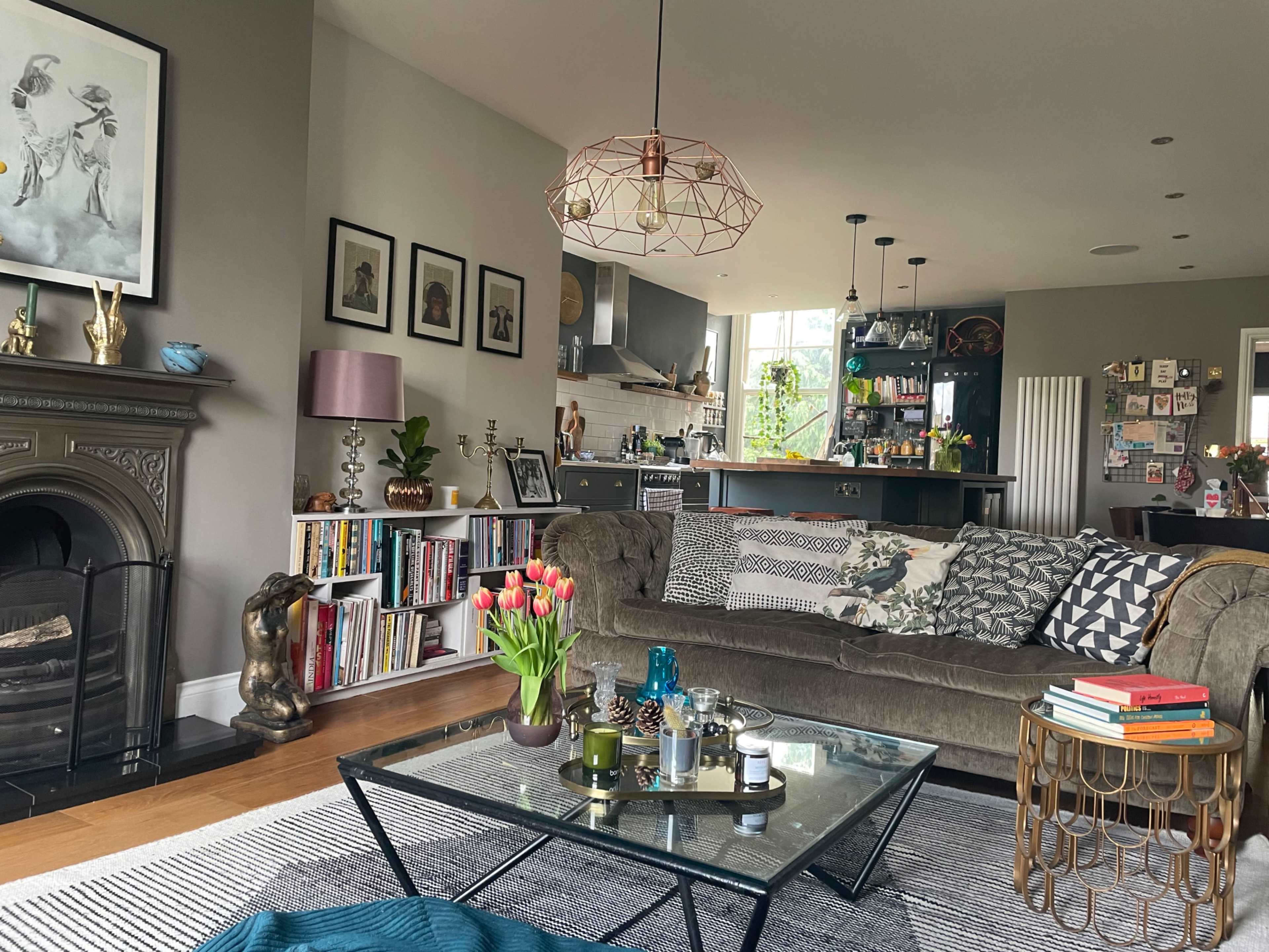 A stylish living room with a brown sofa, a glass coffee table, bookshelves, and colorful decor, leading into a modern kitchen area.