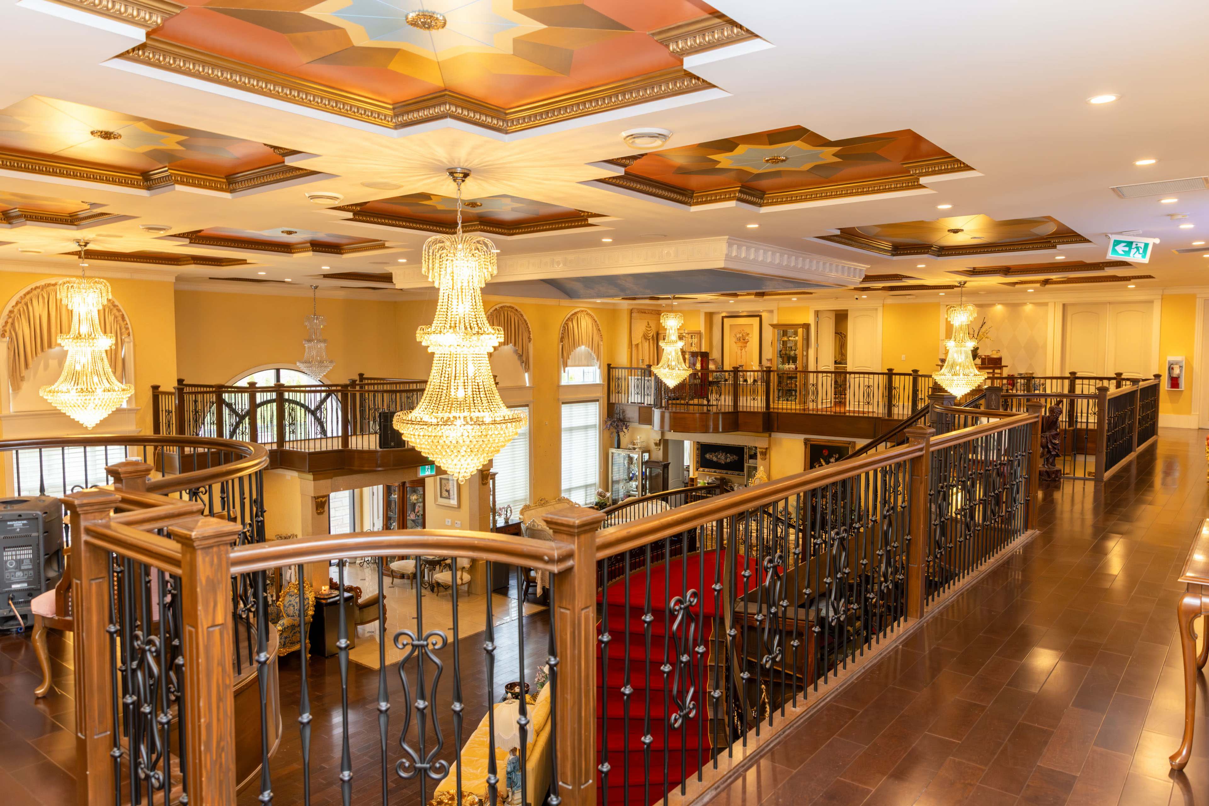 The image shows an elegantly designed interior of a spacious two-story lobby with ornate ceilings, chandeliers, and a wooden railing overlooking a red-carpeted staircase.