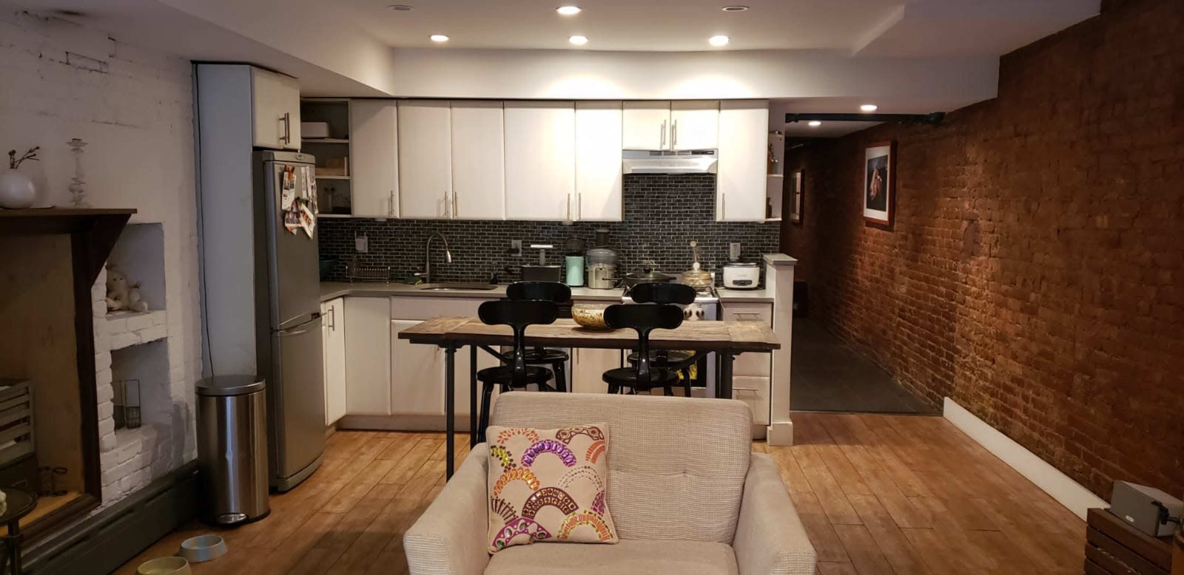 A modern kitchen and living area with a gray couch, two black bar stools at a kitchen island, and exposed brick walls.