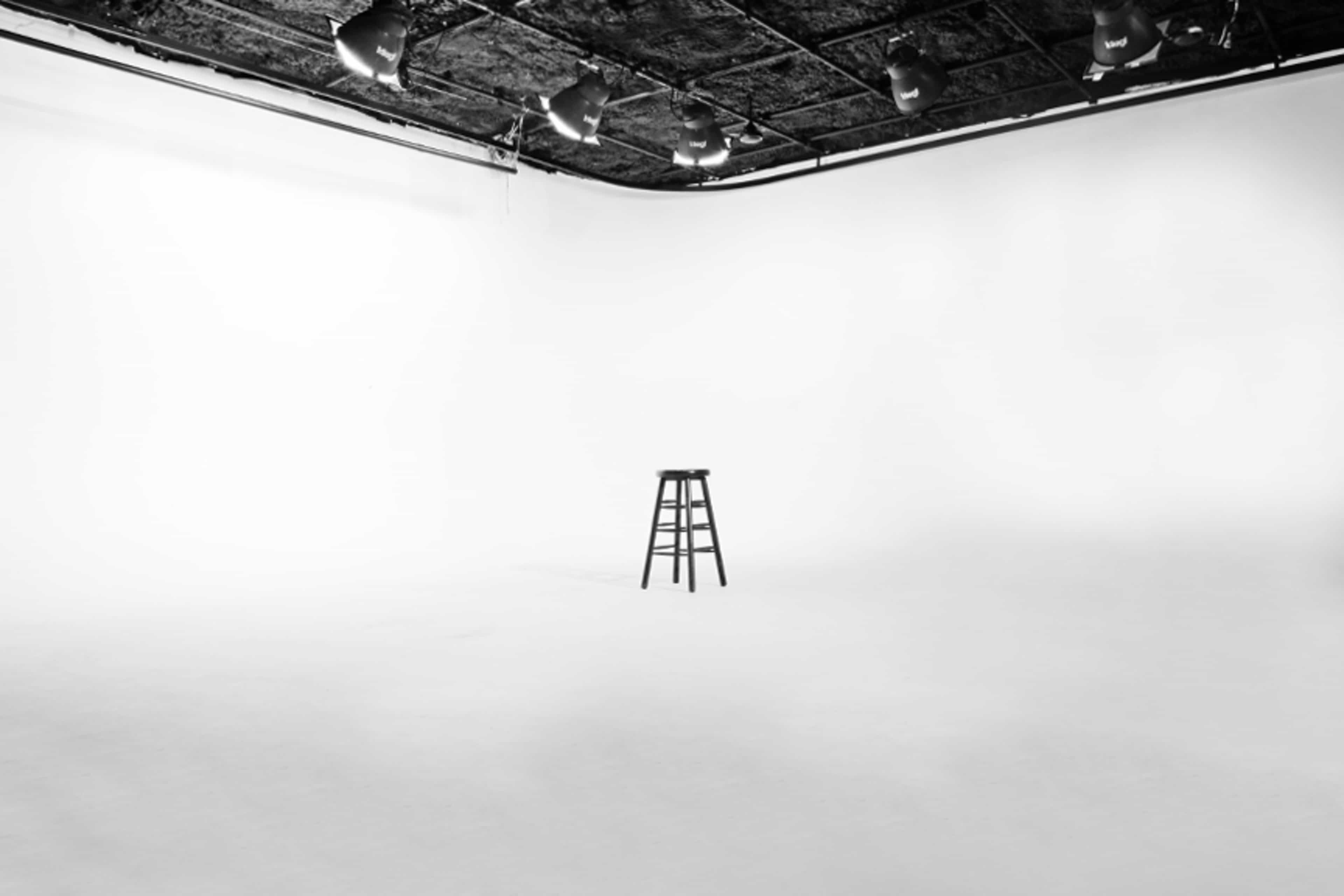 A black stool stands alone in a brightly lit, empty studio with white walls.