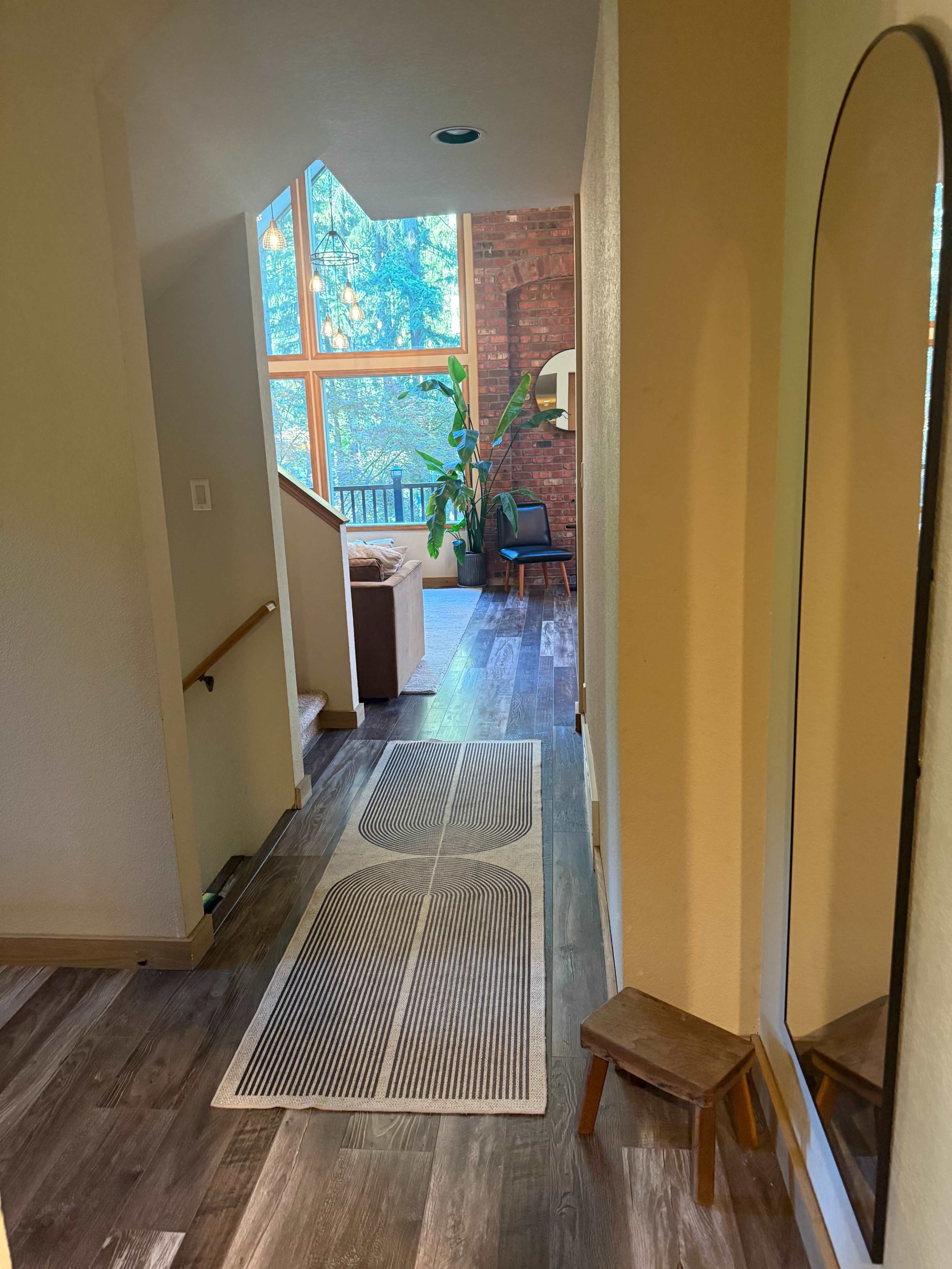 A long hallway leads to a living area with large windows, a sofa, and a chair, featuring a mirror and a small stool on the left.