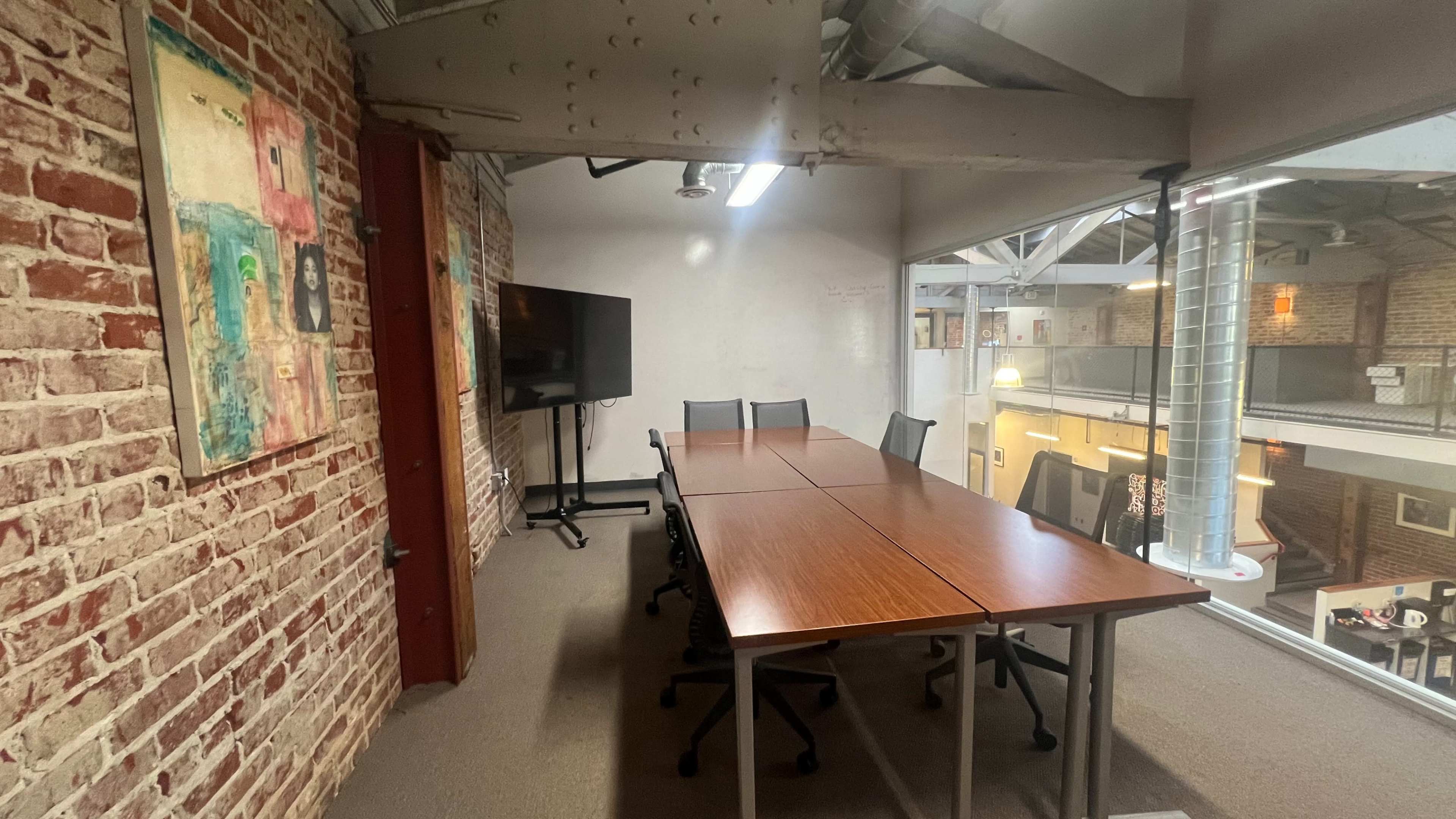 A conference room features a long wooden table surrounded by several chairs, with exposed brick walls and large windows overlooking an open office space.