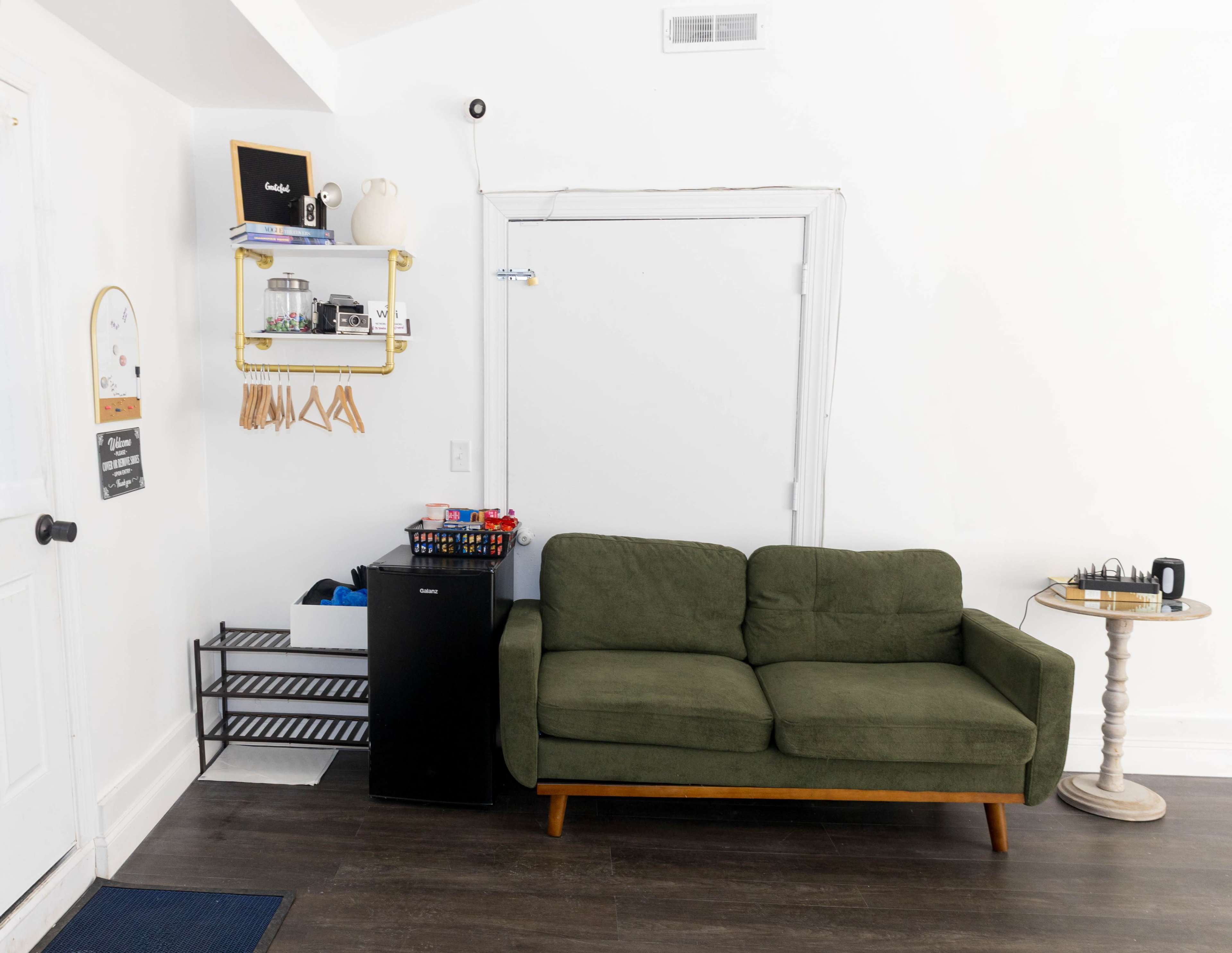 The image shows a small living area featuring a green sofa, a black mini refrigerator, a shelf with storage items, and a small round table.