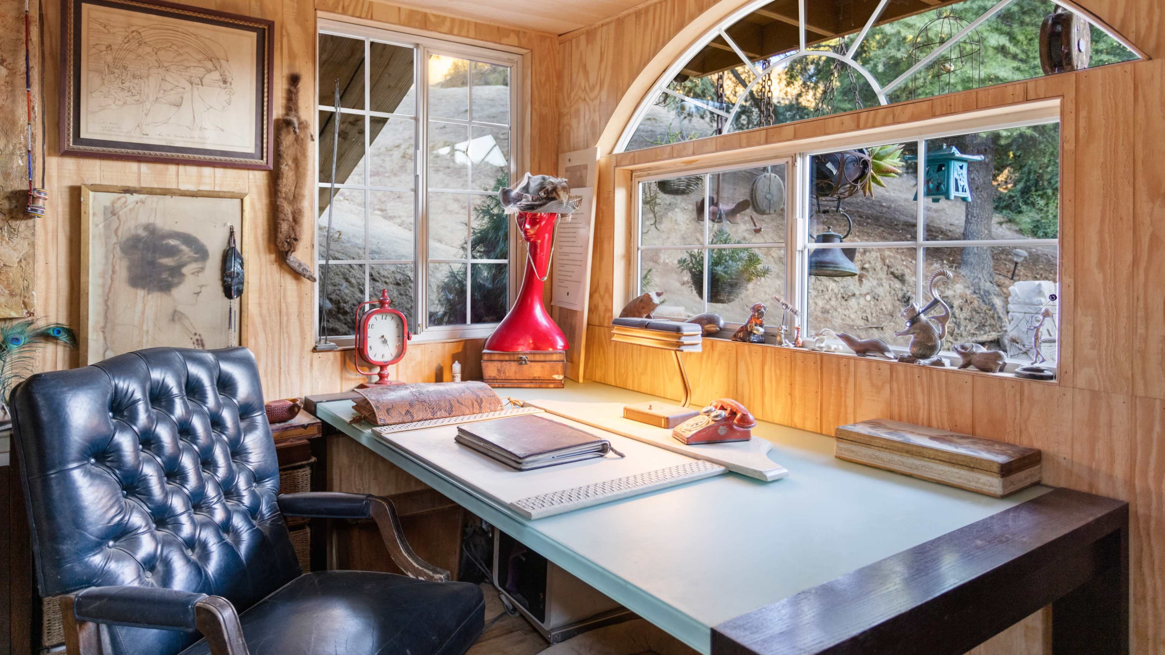 The image shows a wooden desk with a blue surface, an antique clock, and various items on it, positioned next to large windows with a view of greenery outside.