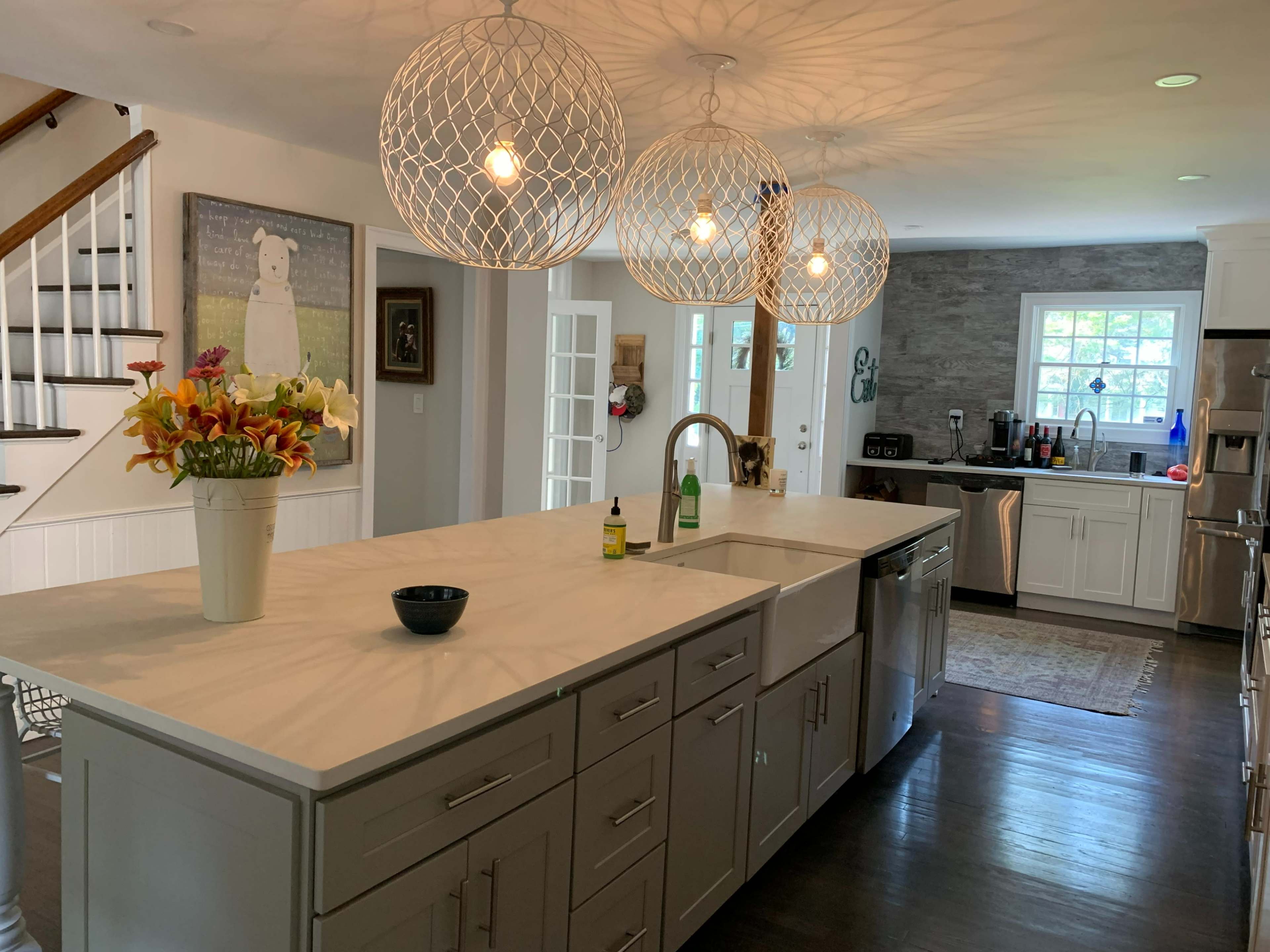 The image shows a modern kitchen featuring a large island with a marble countertop, three pendant lights overhead, and a vase of flowers on the island.