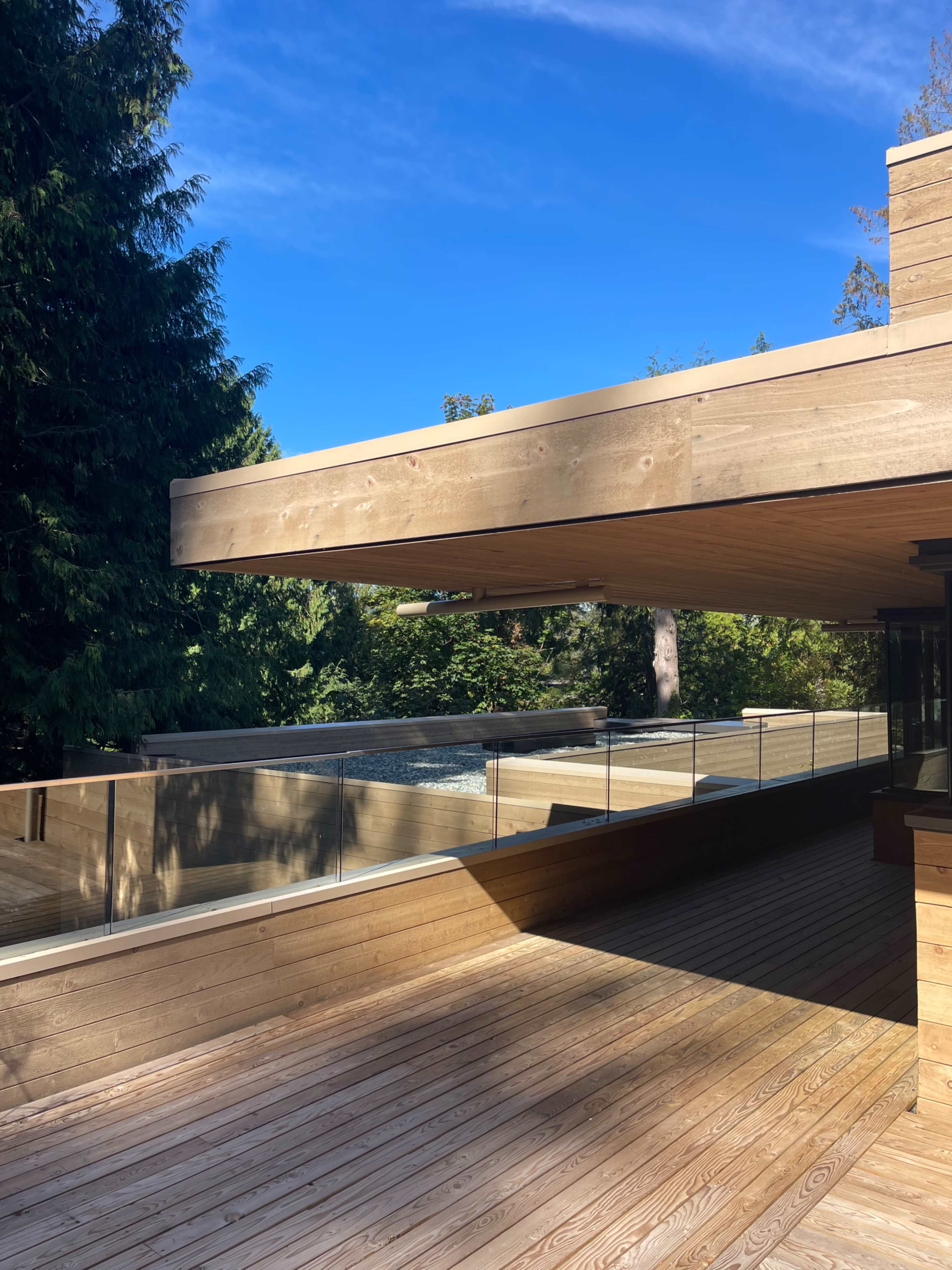 The image shows a modern wooden deck with a glass railing, overlooking a pool surrounded by trees under a clear blue sky.