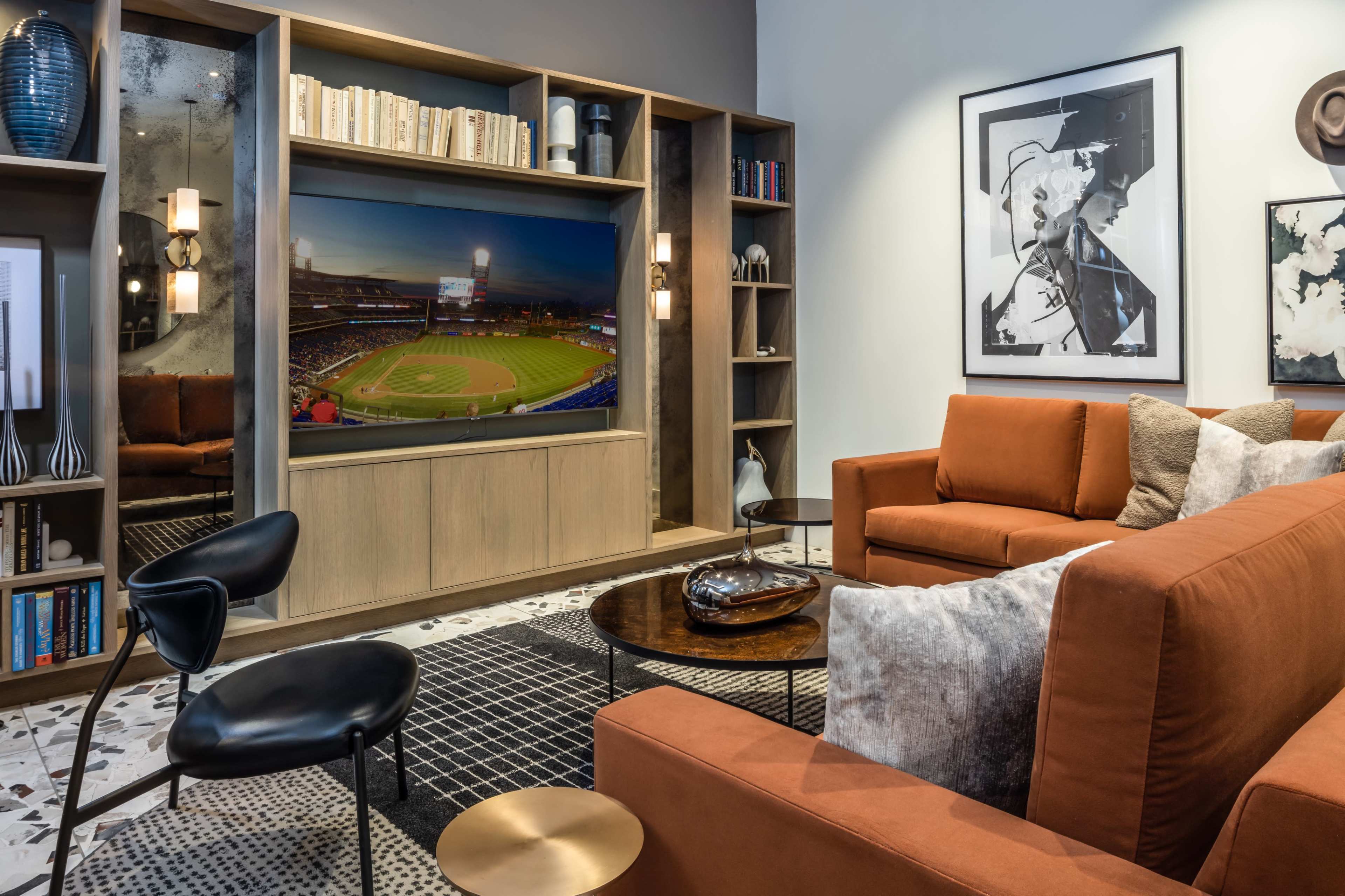A modern living room with a sectional sofa, a round coffee table, and a large television displaying a baseball game, surrounded by bookshelves and artwork.