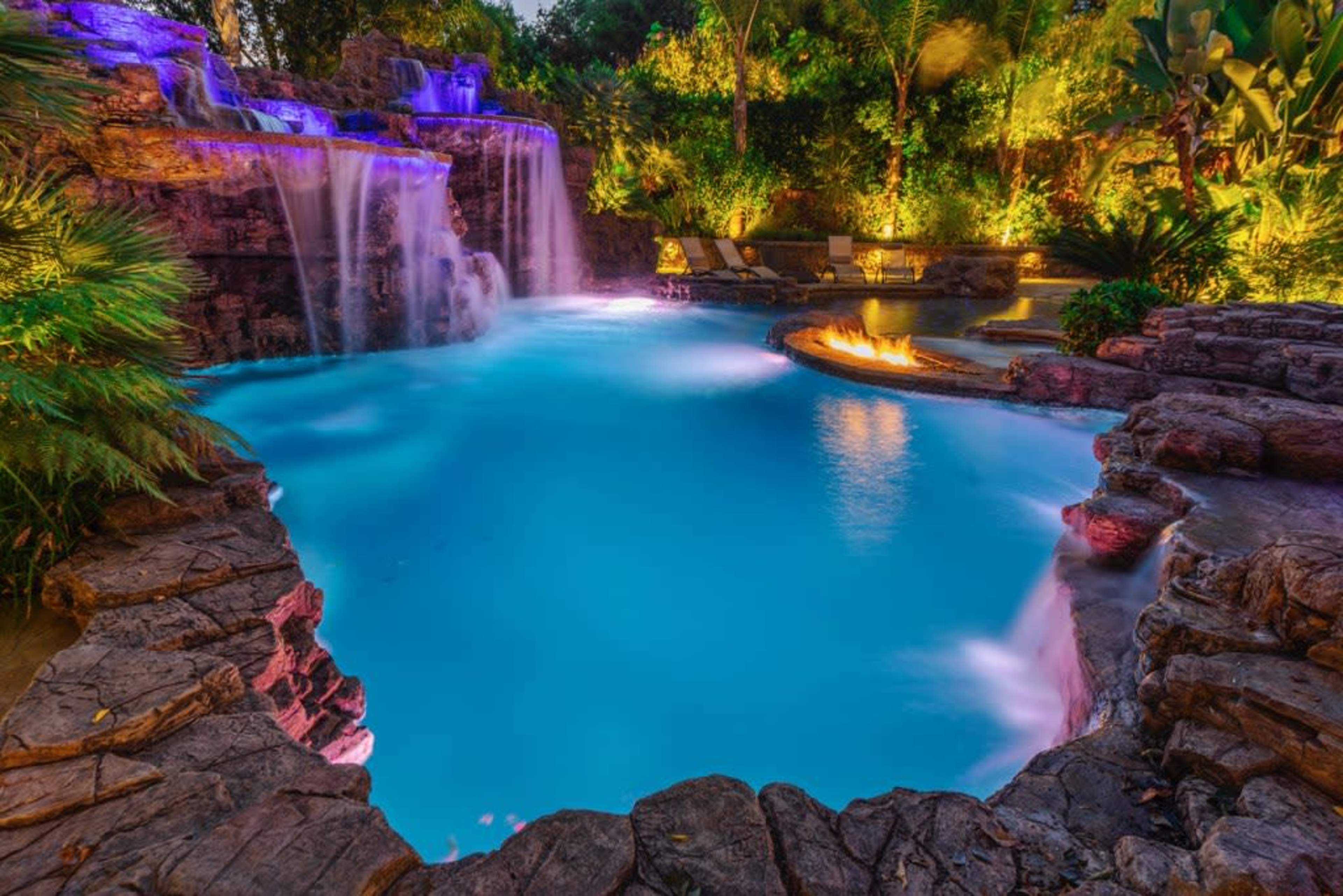 The image shows a beautifully lit swimming pool with a waterfall feature, surrounded by lush greenery and a fire pit.