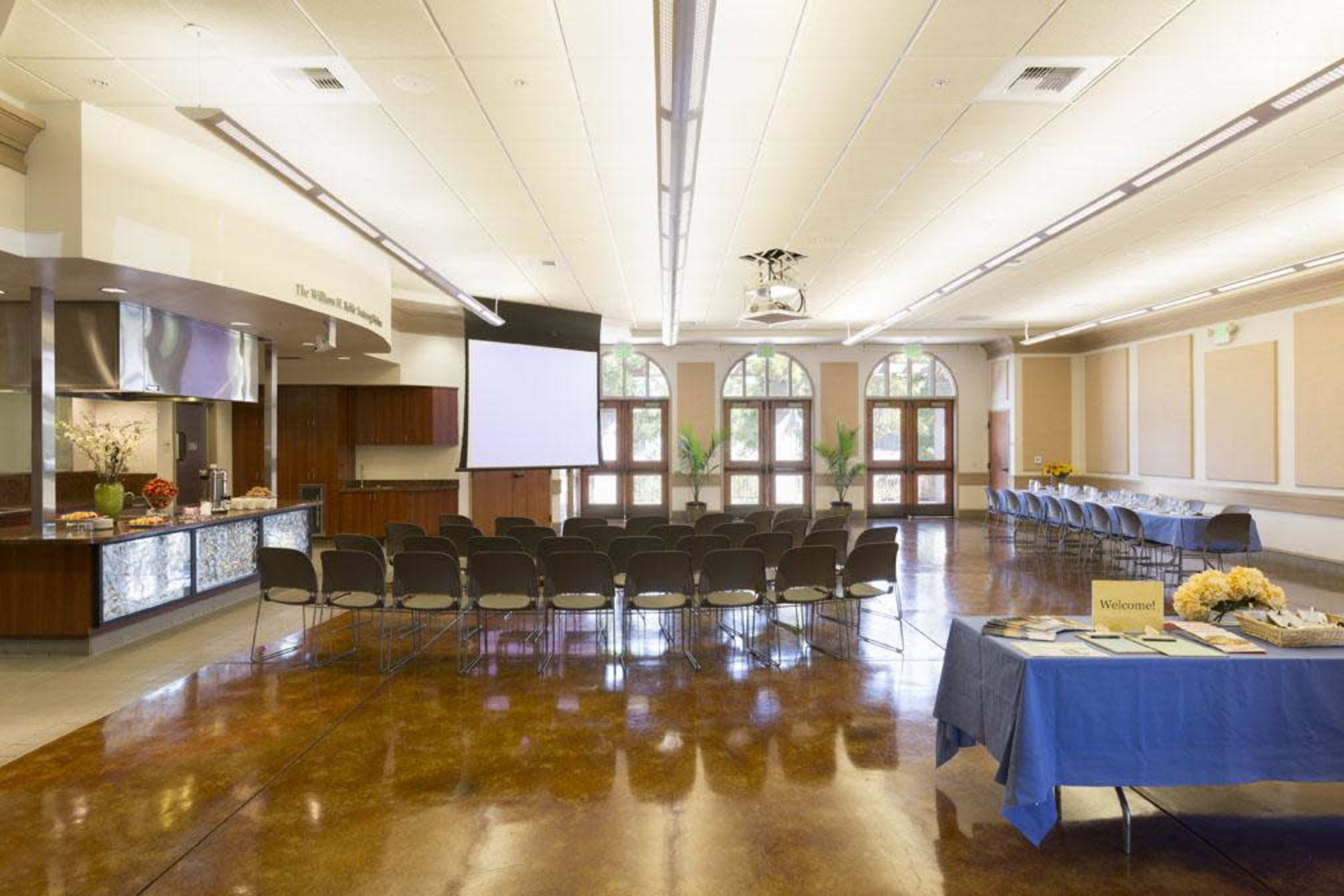 A spacious event room set up with rows of chairs facing a projection screen, with a refreshments table on one side.