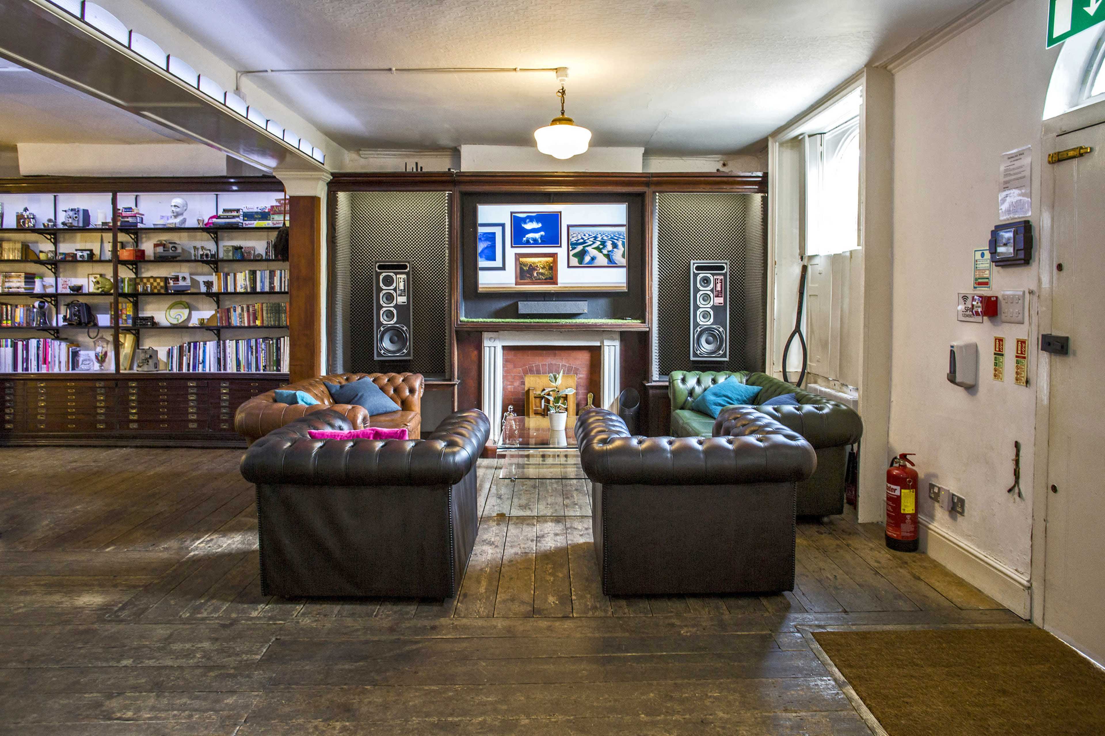 The image shows a room with a seating area featuring leather sofas arranged around a fireplace and a large screen mounted on the wall, with bookshelves in the background.