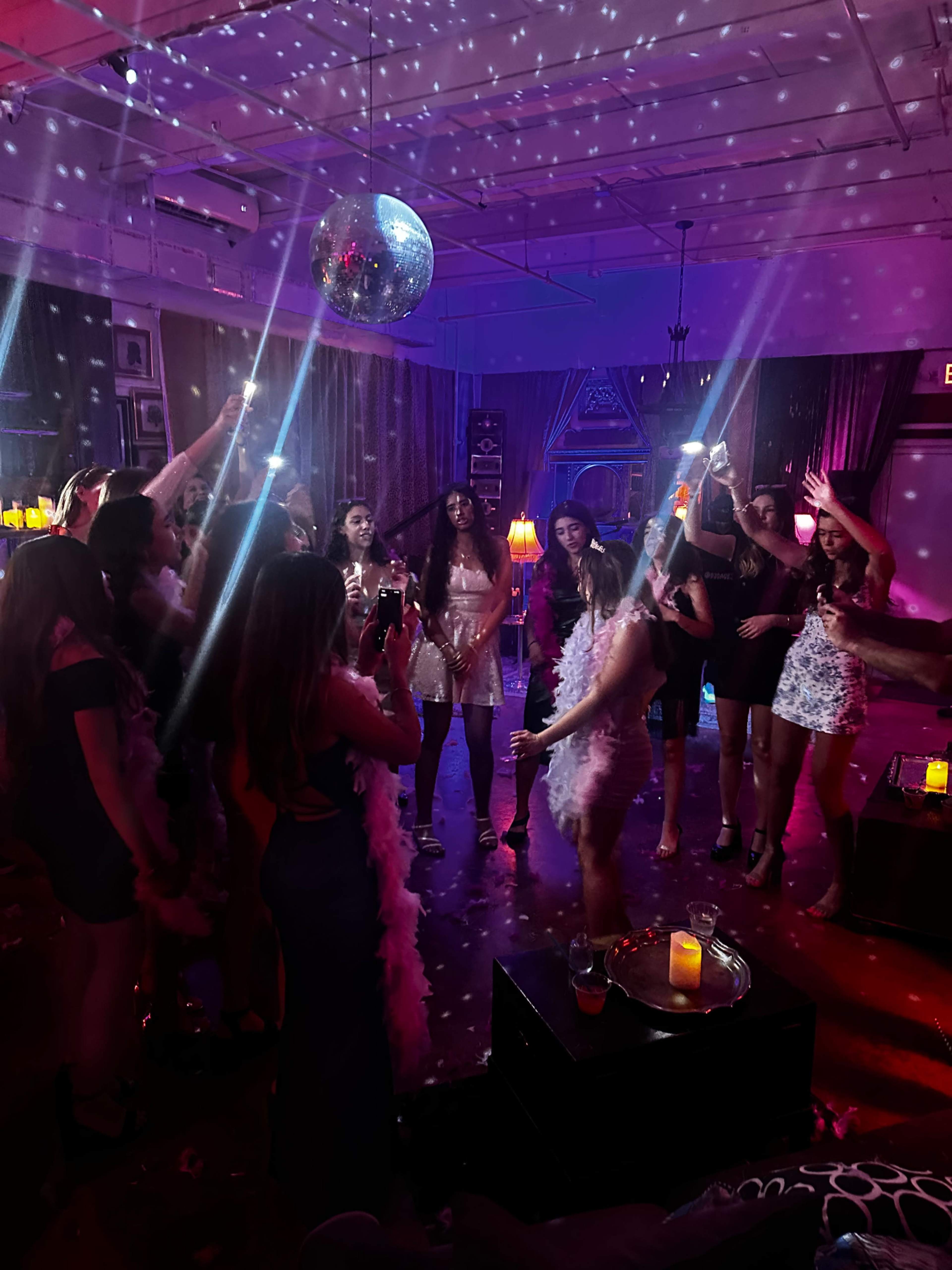 A group of people celebrate in a dimly lit room adorned with a disco ball and colorful lights while holding drinks and dancing.