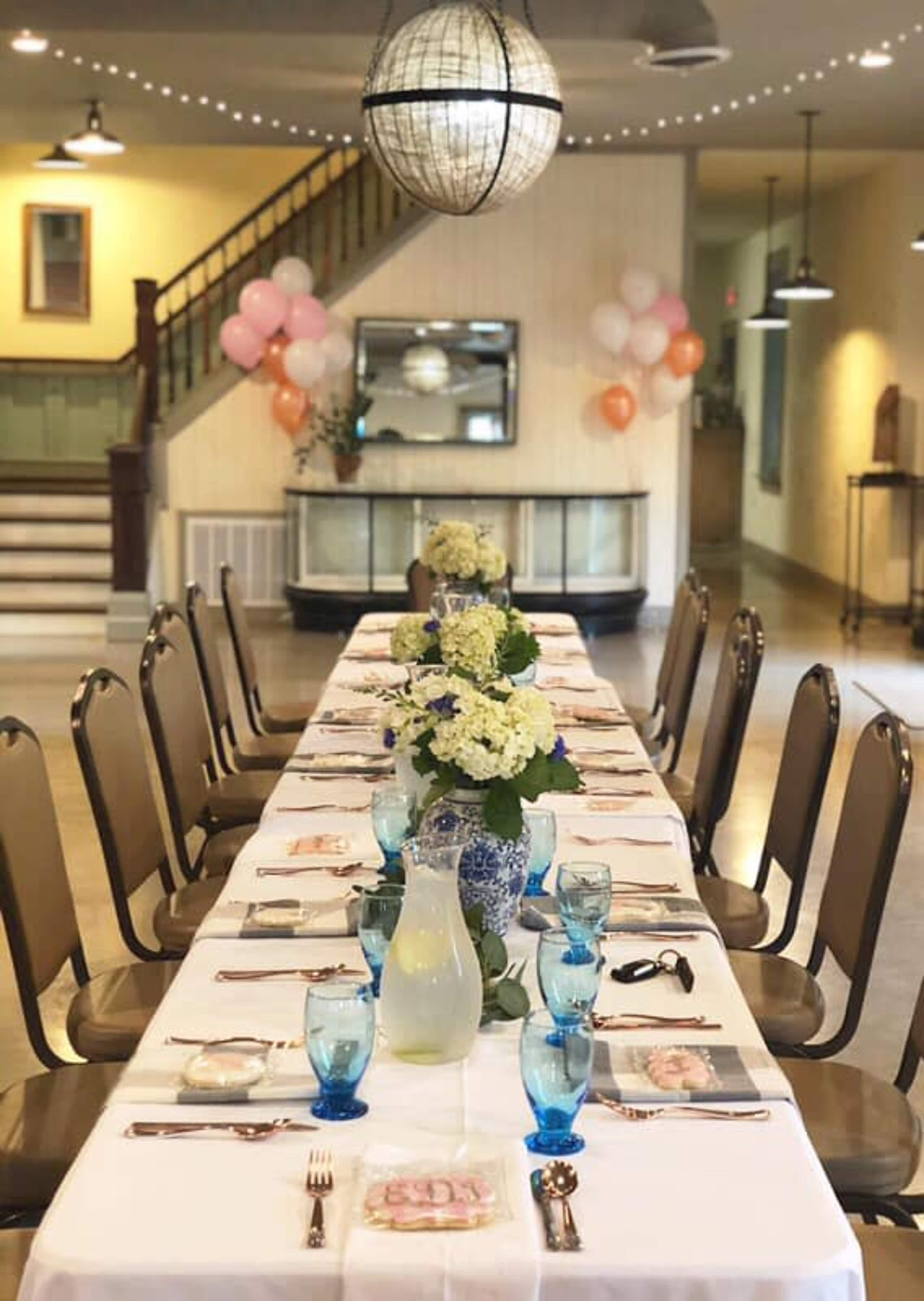 A long dining table is set with blue glassware and floral centerpieces, surrounded by metal chairs in a decorated banquet space.