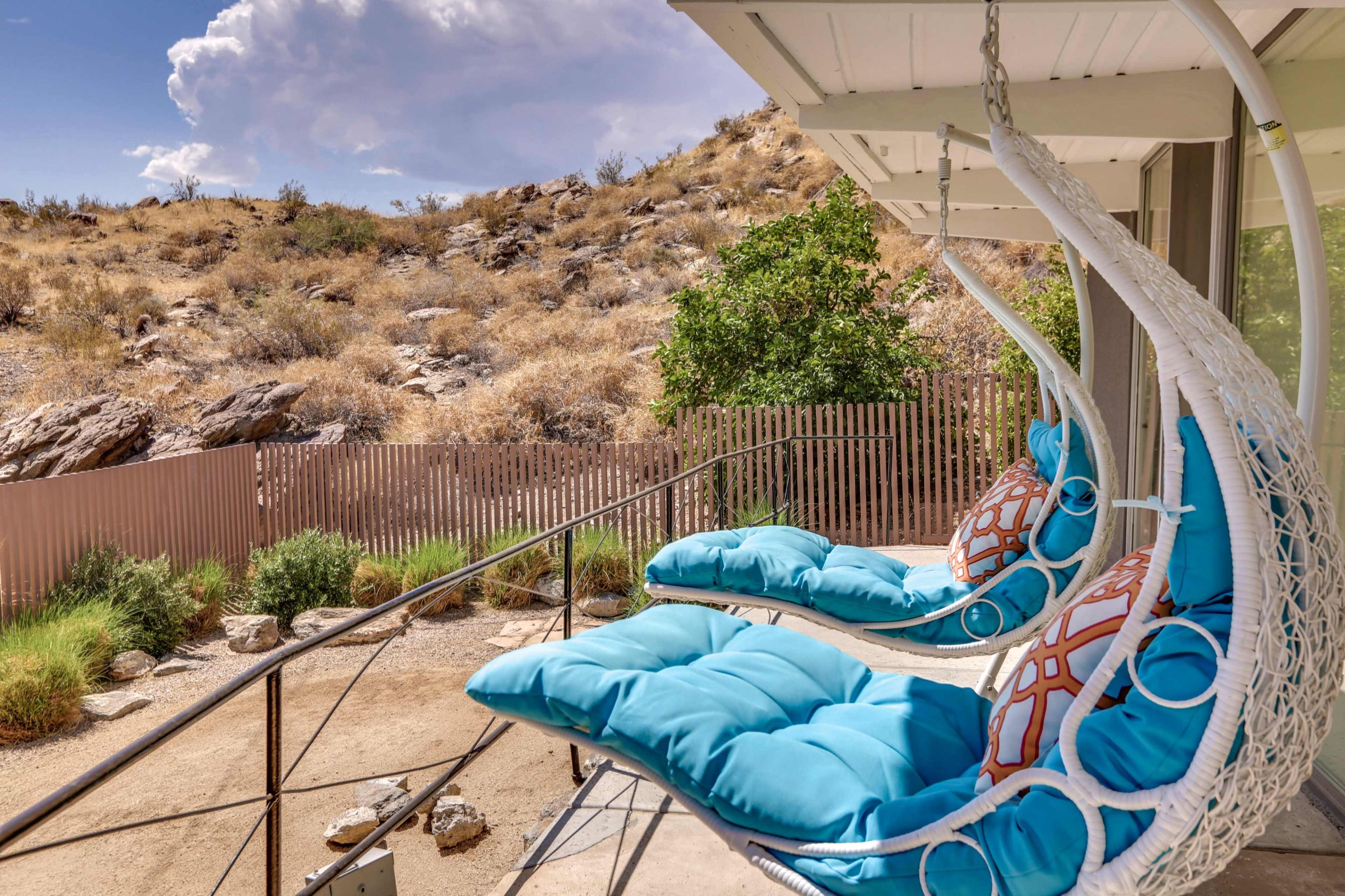 A pair of hanging chairs with blue cushions is positioned on a balcony overlooking a hillside covered with dry vegetation.