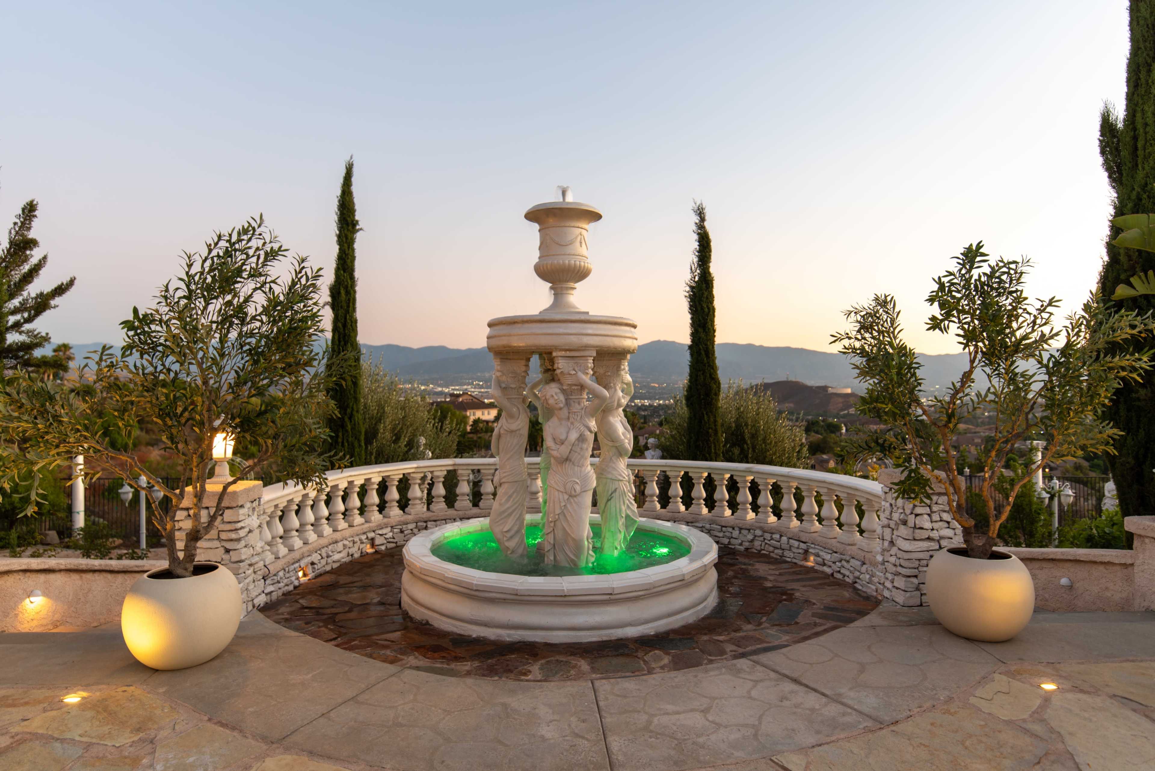 The image shows a stone terrace with a decorative fountain featuring three female figures, surrounded by olive trees and distant mountains under a fading light.