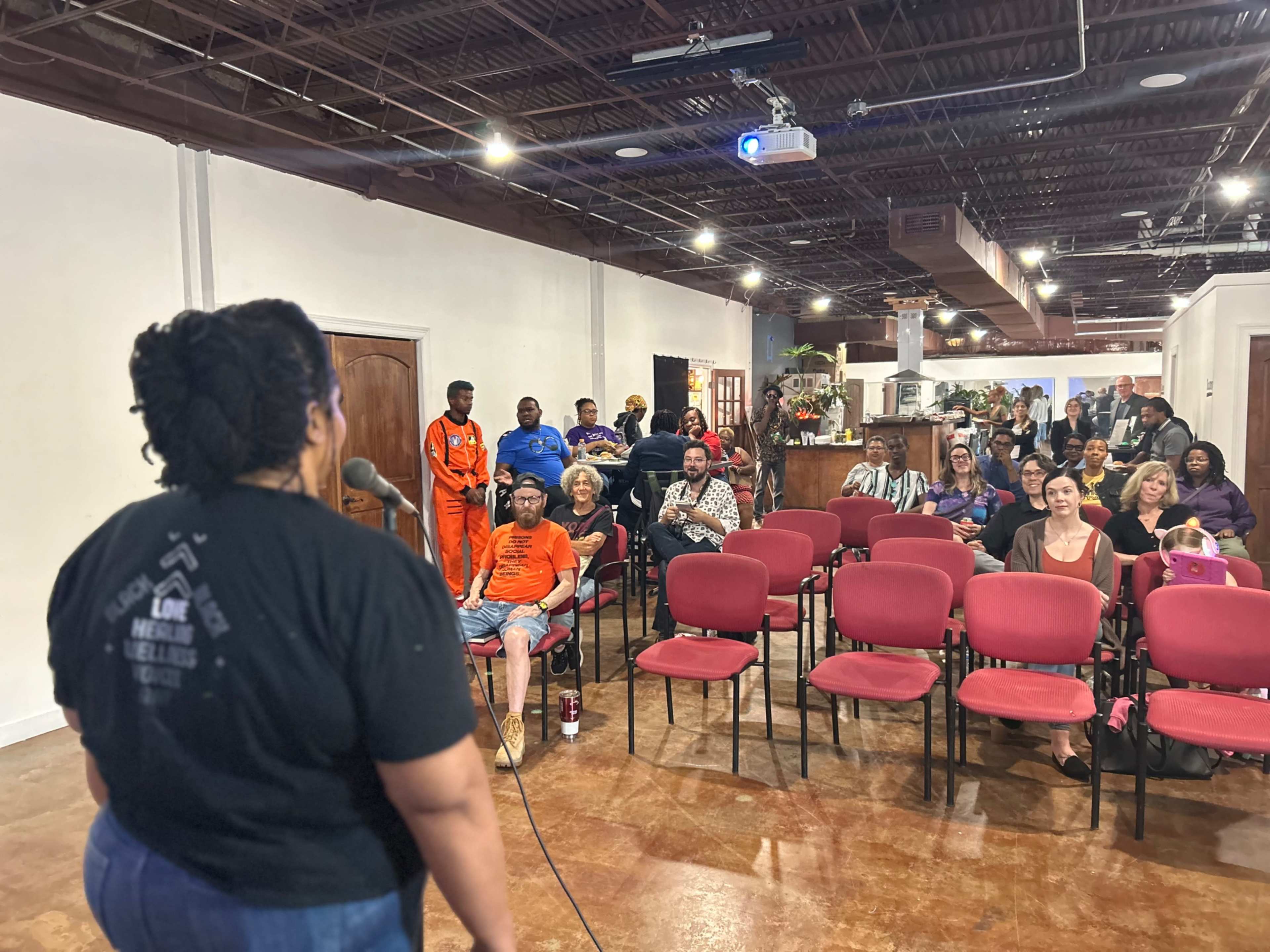 A speaker stands at a microphone in front of an audience seated in chairs at a community event venue.