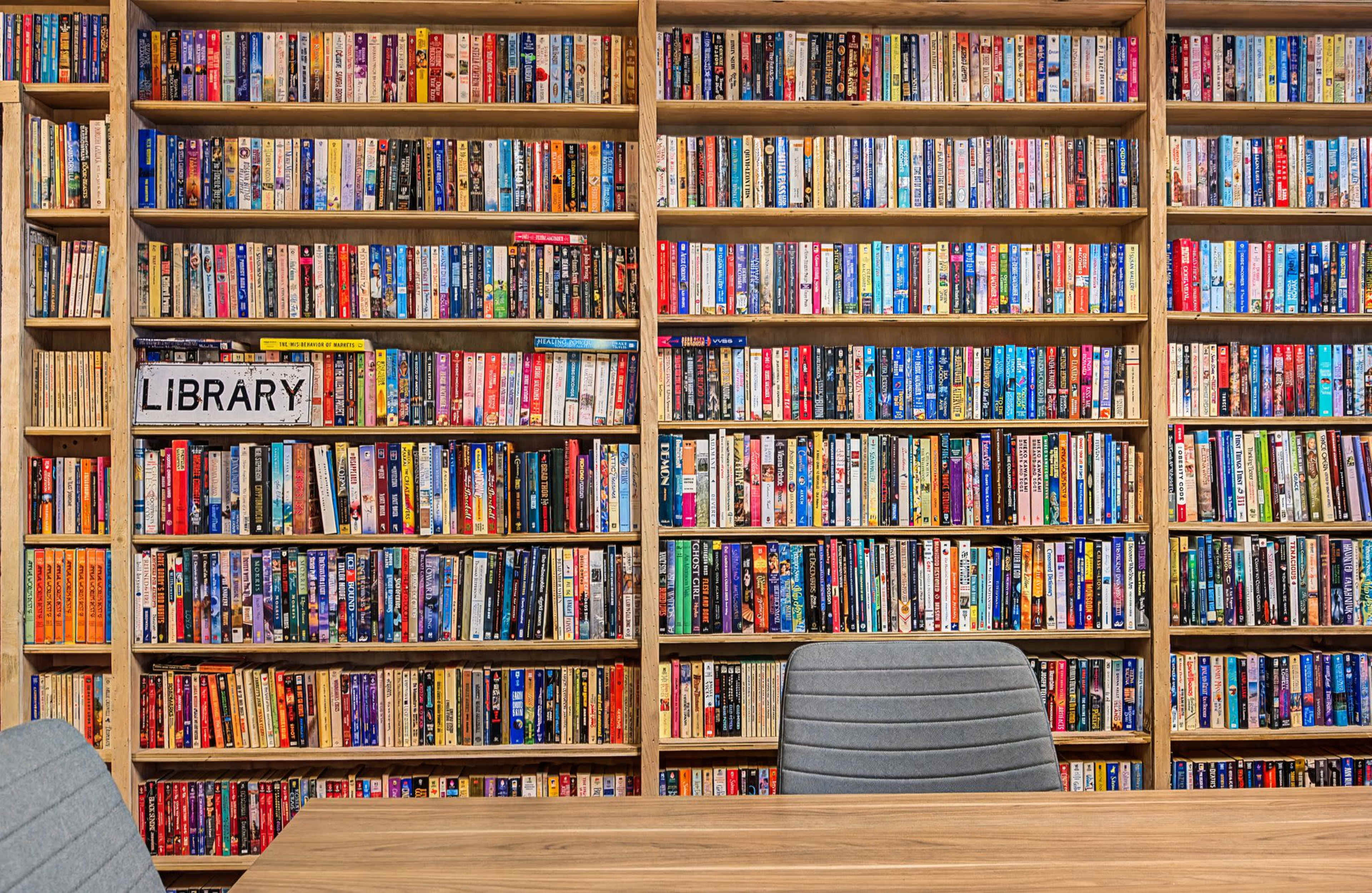 A wall of shelves filled with a wide variety of books stands behind a wooden table with two gray chairs.