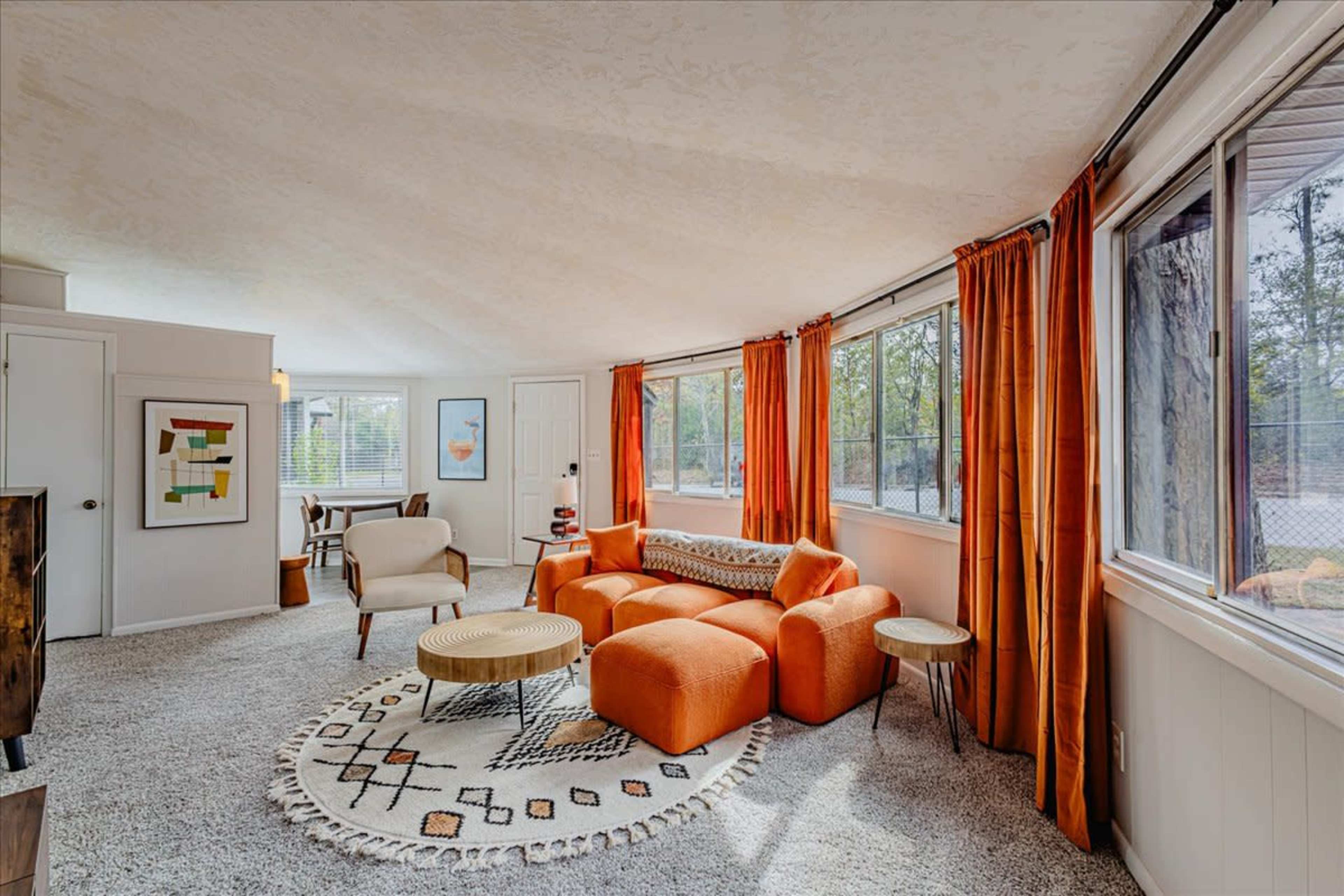 A cozy living room featuring orange furniture, large windows allowing natural light, and a patterned rug on a carpeted floor.
