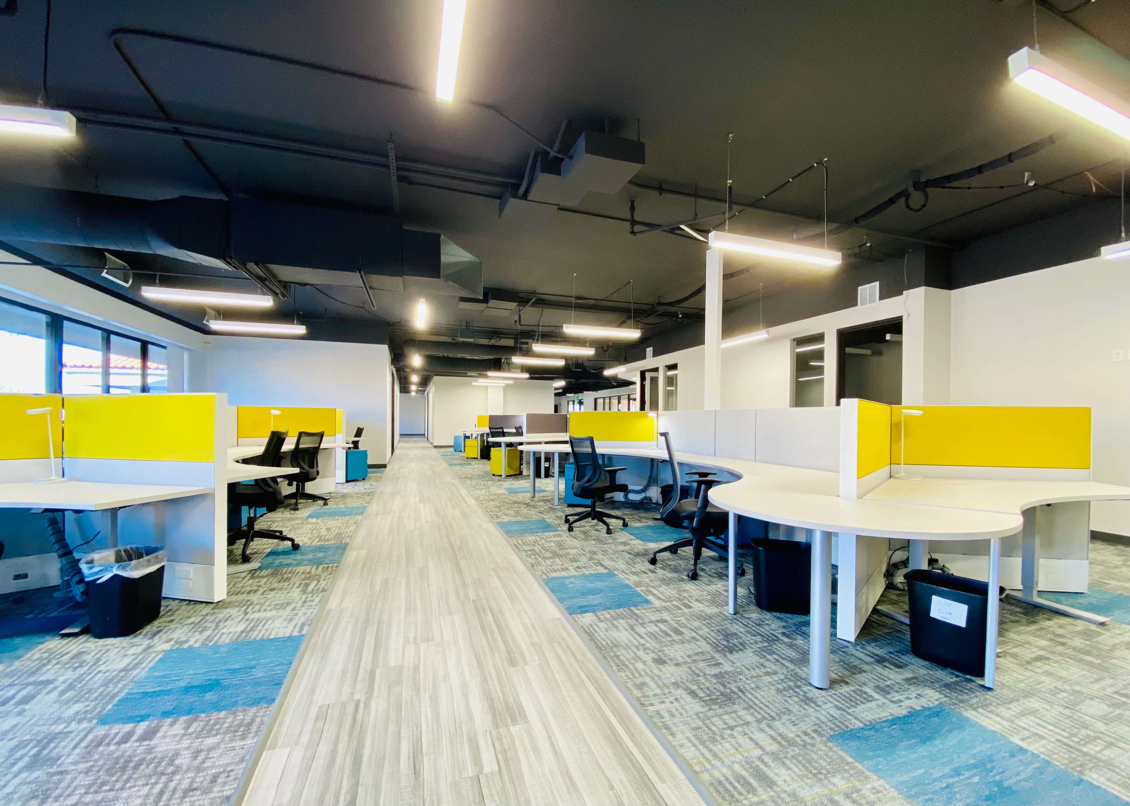 A modern office space with multiple workstations arranged along a central corridor, accented by yellow dividers and carpeted flooring.