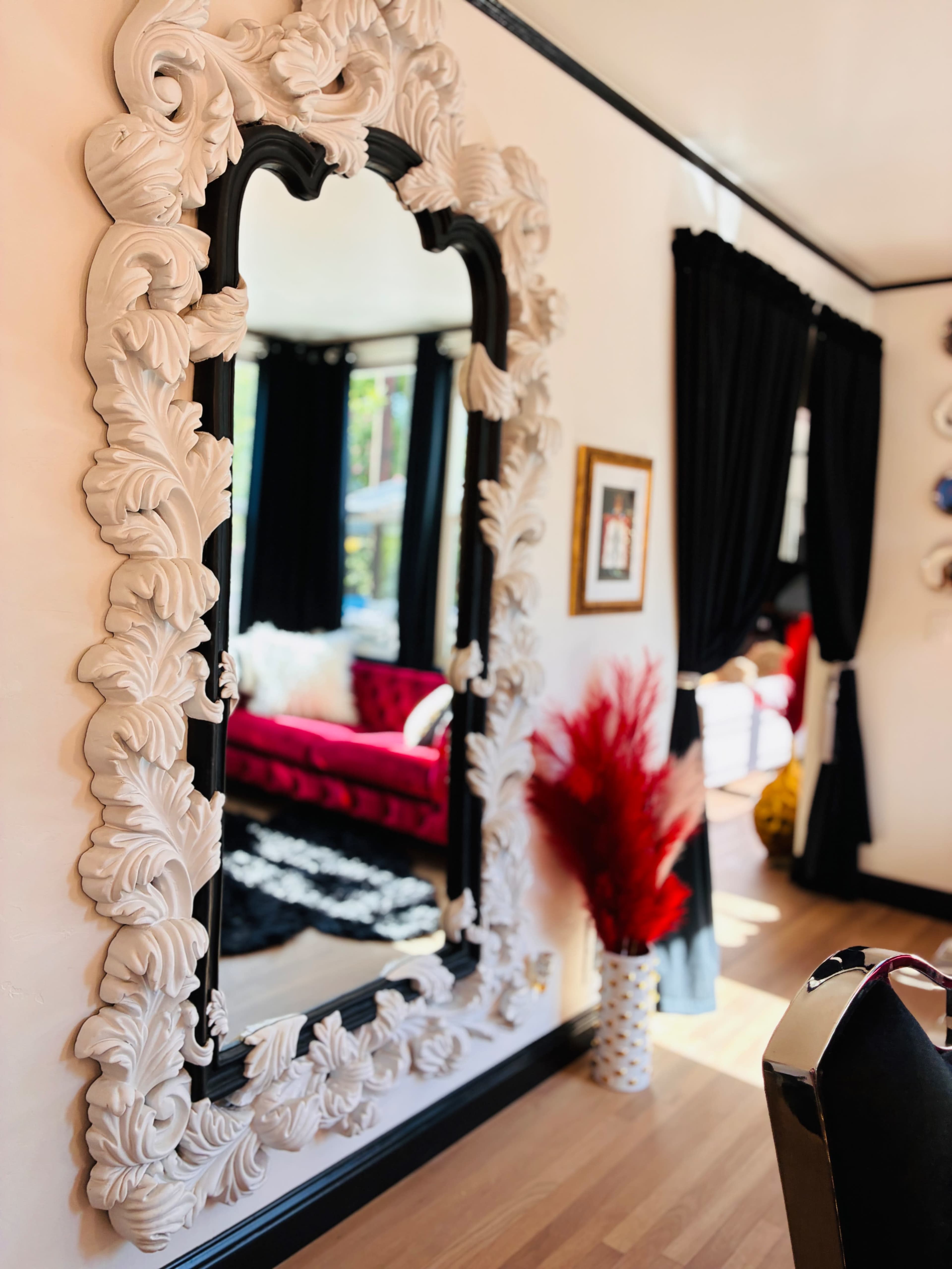 The image shows a large ornate mirror on a wall, reflecting a room with a red sofa, black curtains, and decorative plants.