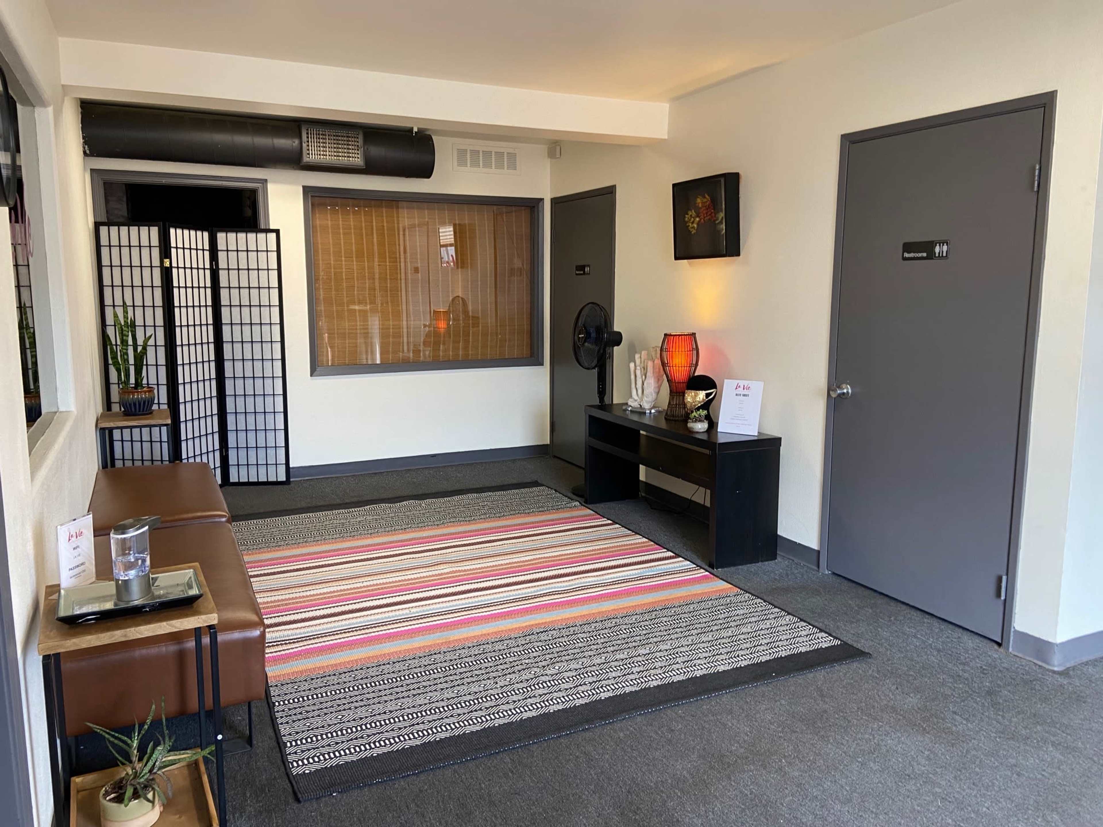 A waiting area features a striped rug, a low wooden bench, a dark side table, and a partition, with plants and decorative items enhancing the space.