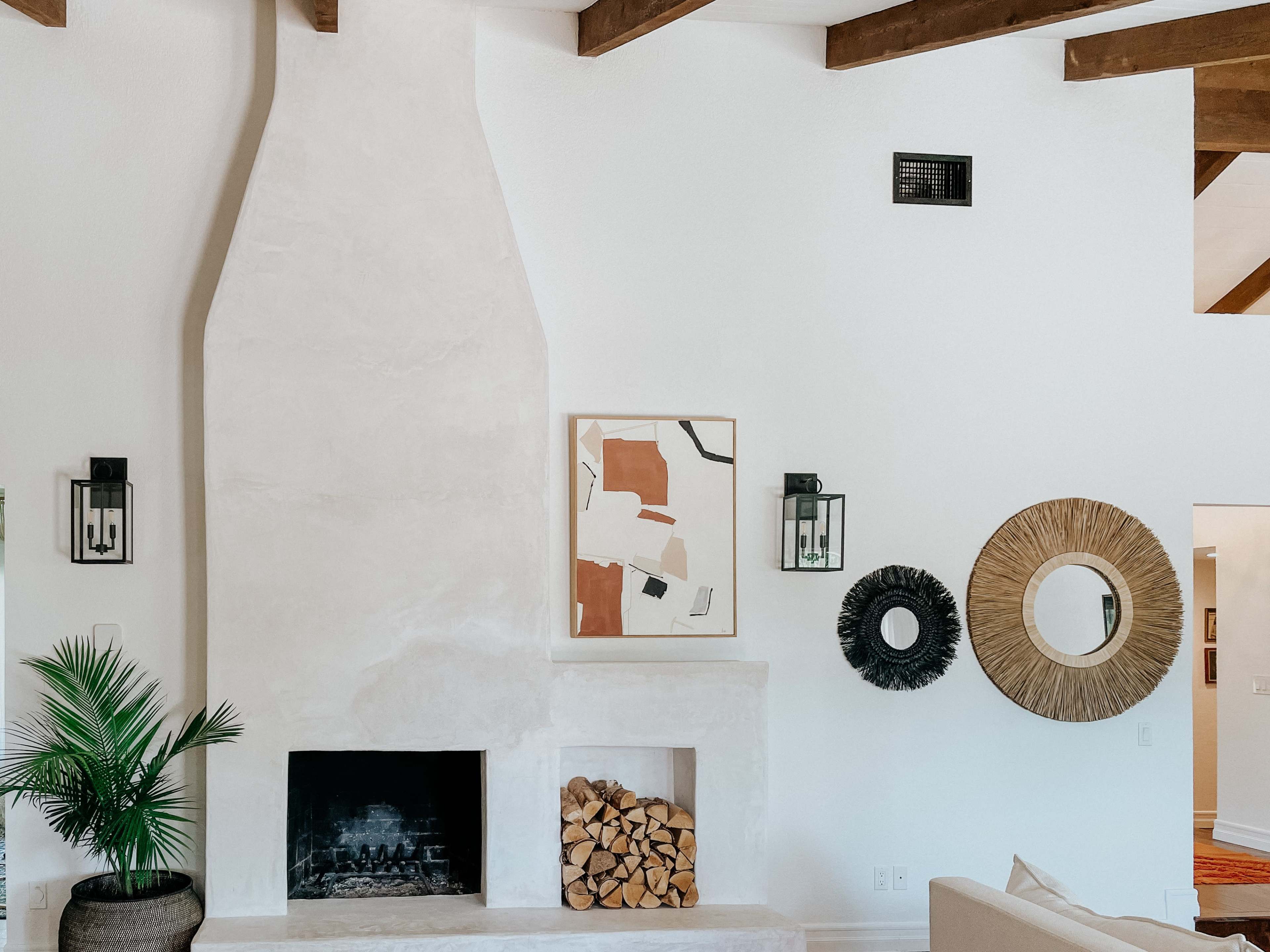 A living room featuring a white plaster fireplace, stacked firewood, decorative mirrors, and framed artwork on the wall, with wooden beams visible in the ceiling.