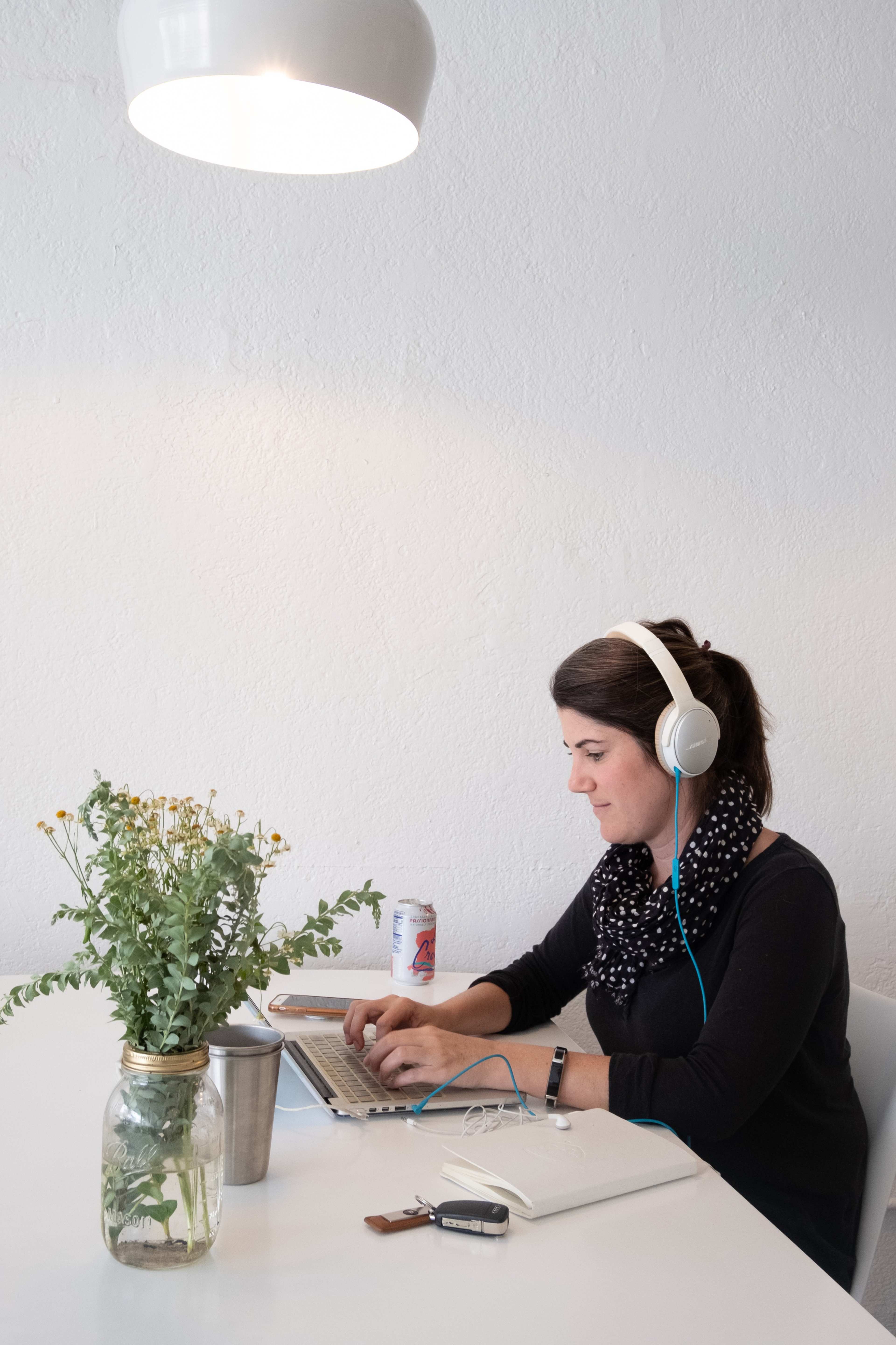A woman wearing headphones sits at a table with a laptop, a notebook, and a flower arrangement in a jar.