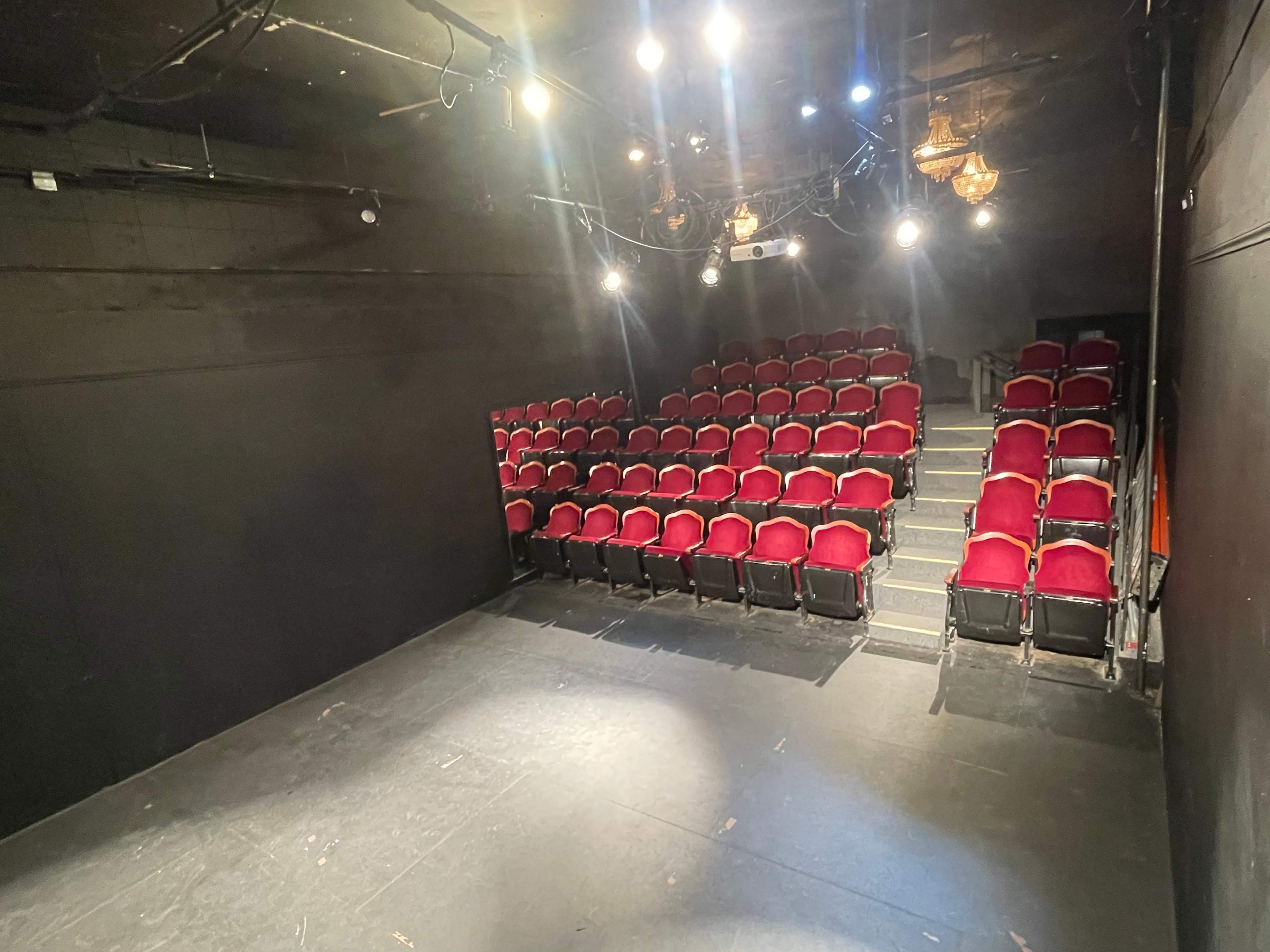 A small theater with red velvet seats arranged in a tiered formation against a black backdrop and bright overhead lights.