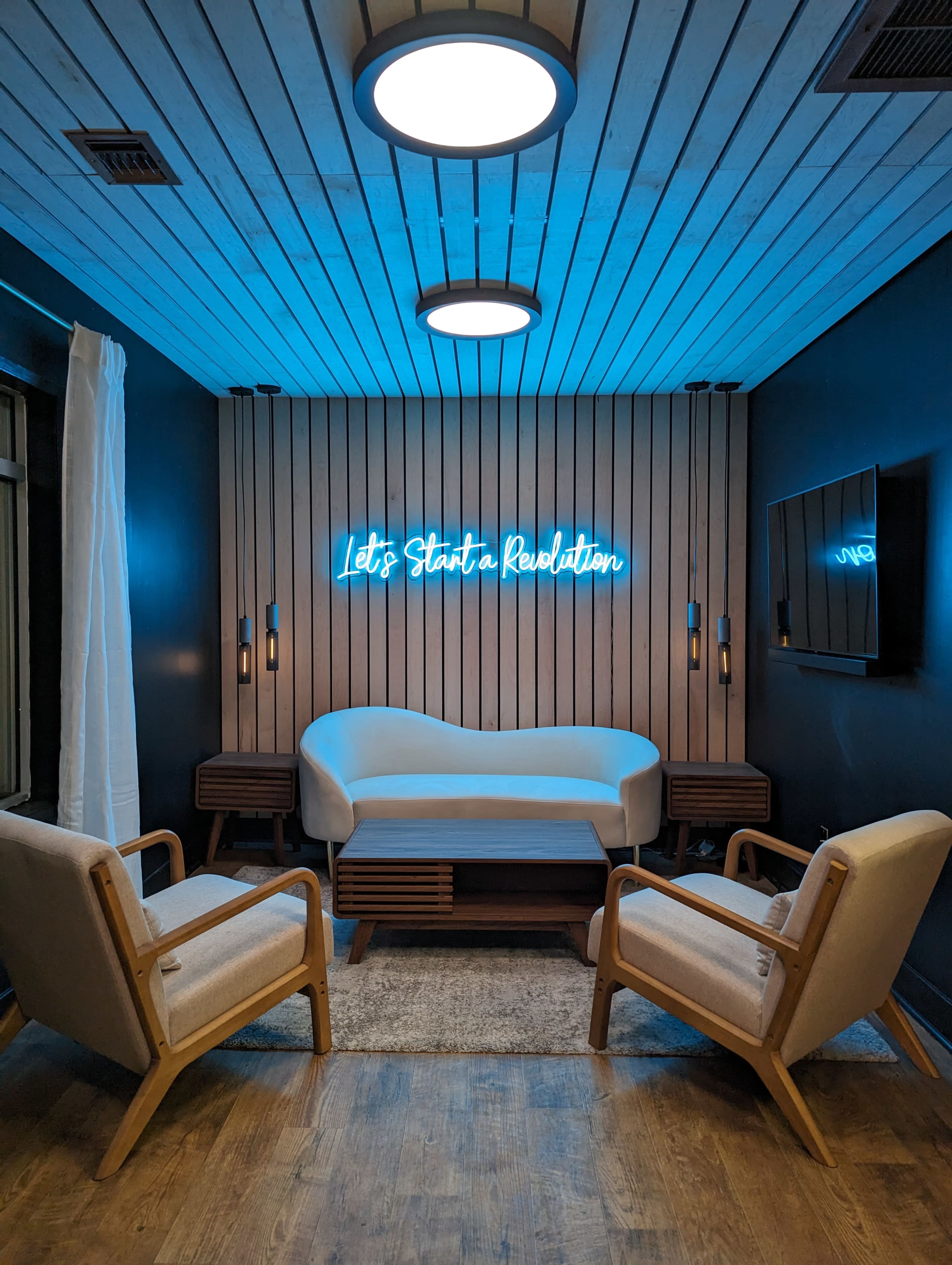 A modern lounge area with a white sofa, two light-colored chairs, a neon sign that reads "Let's Start a Revolution," and a television mounted on the wall.