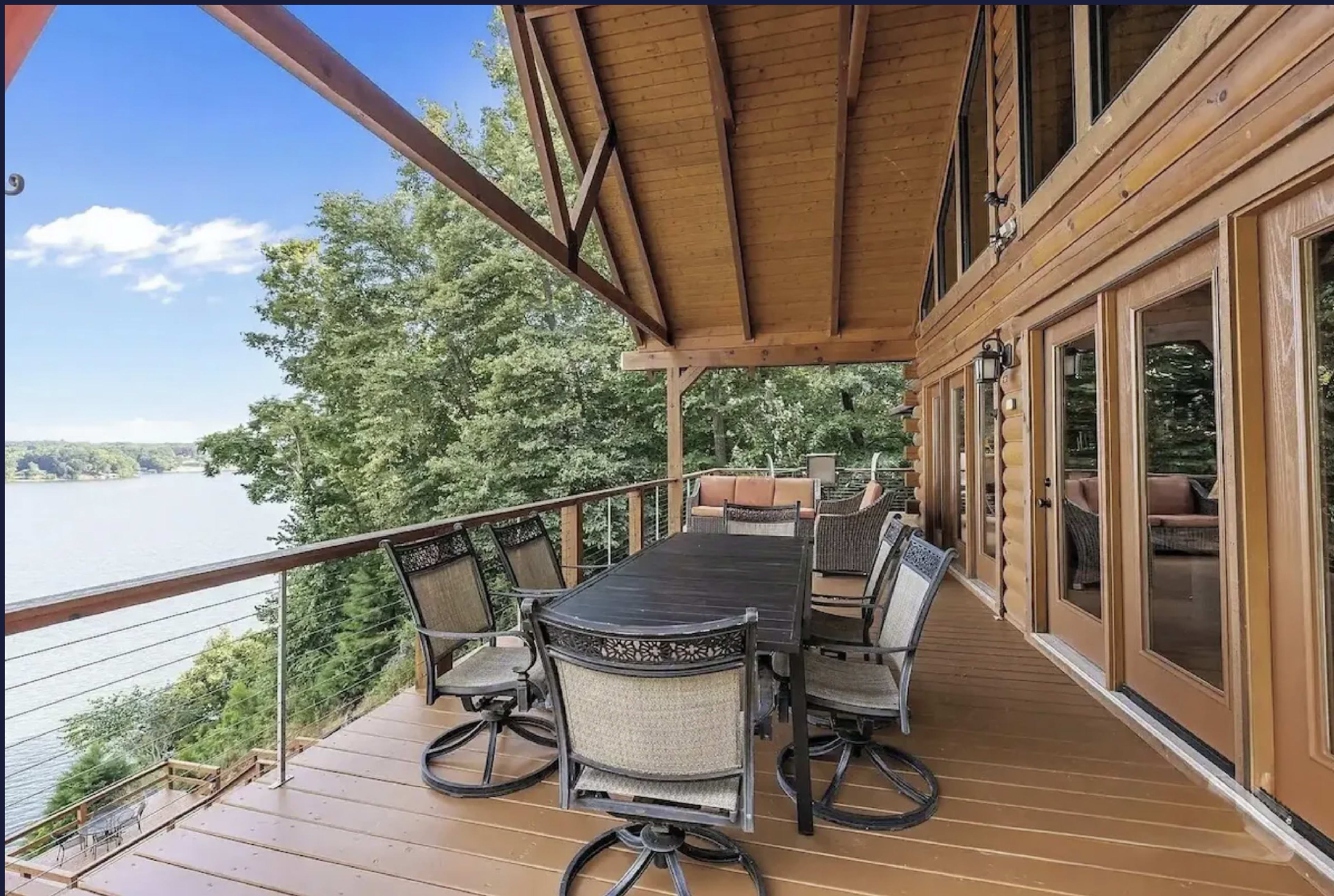 A spacious deck with a large dining table and numerous chairs overlooks a calm lake and surrounding trees.