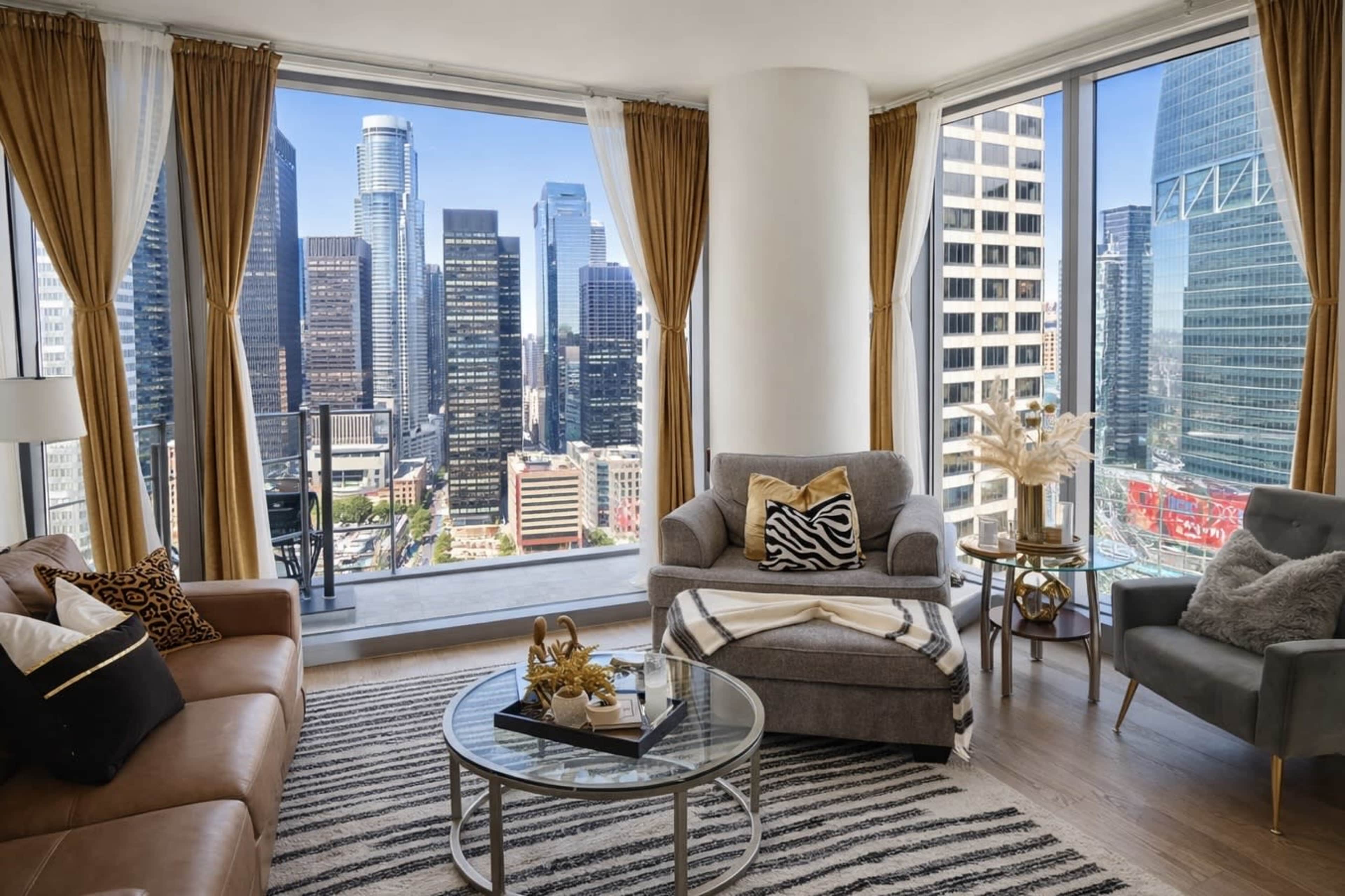 The image shows a modern apartment living room with large windows offering a view of a city skyline, featuring a mix of seating arrangements and decorative elements.