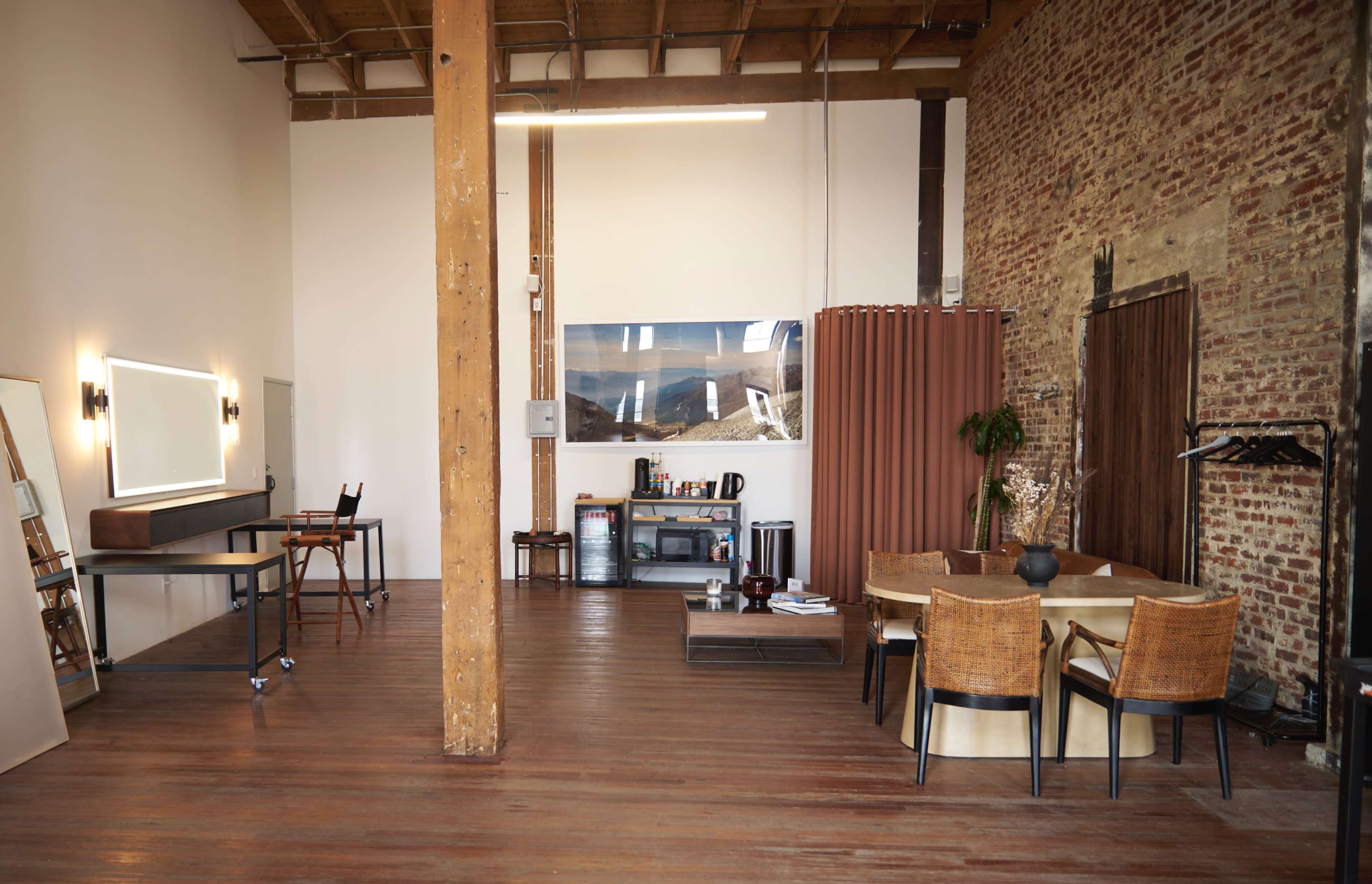 The image shows a spacious, modern interior of a loft with exposed brick walls, wooden beams, and a dining area featuring a round table and chairs.