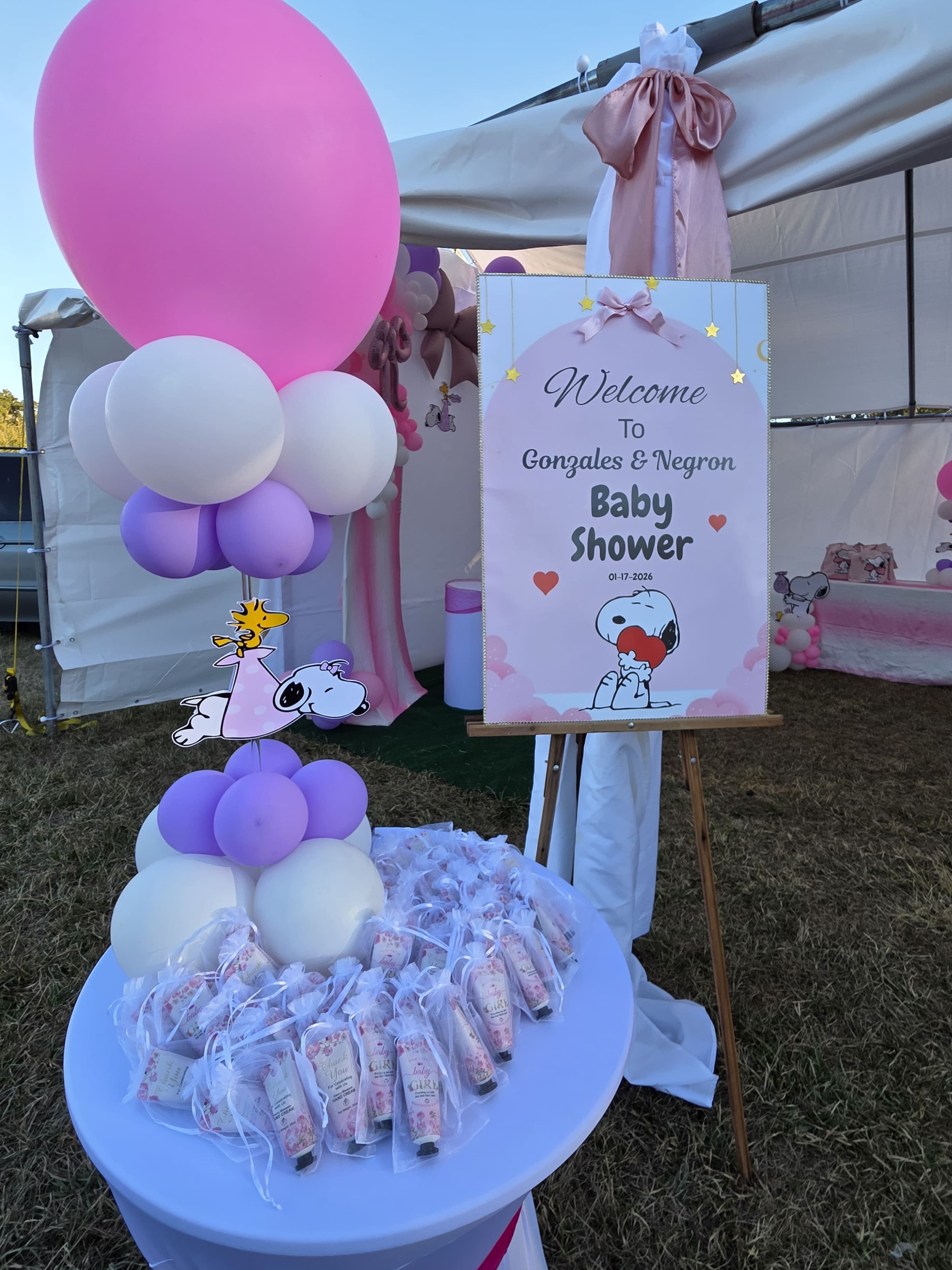 The image shows a decorative setup for a baby shower featuring a welcome sign, a table with favors, and balloons in pink and white.