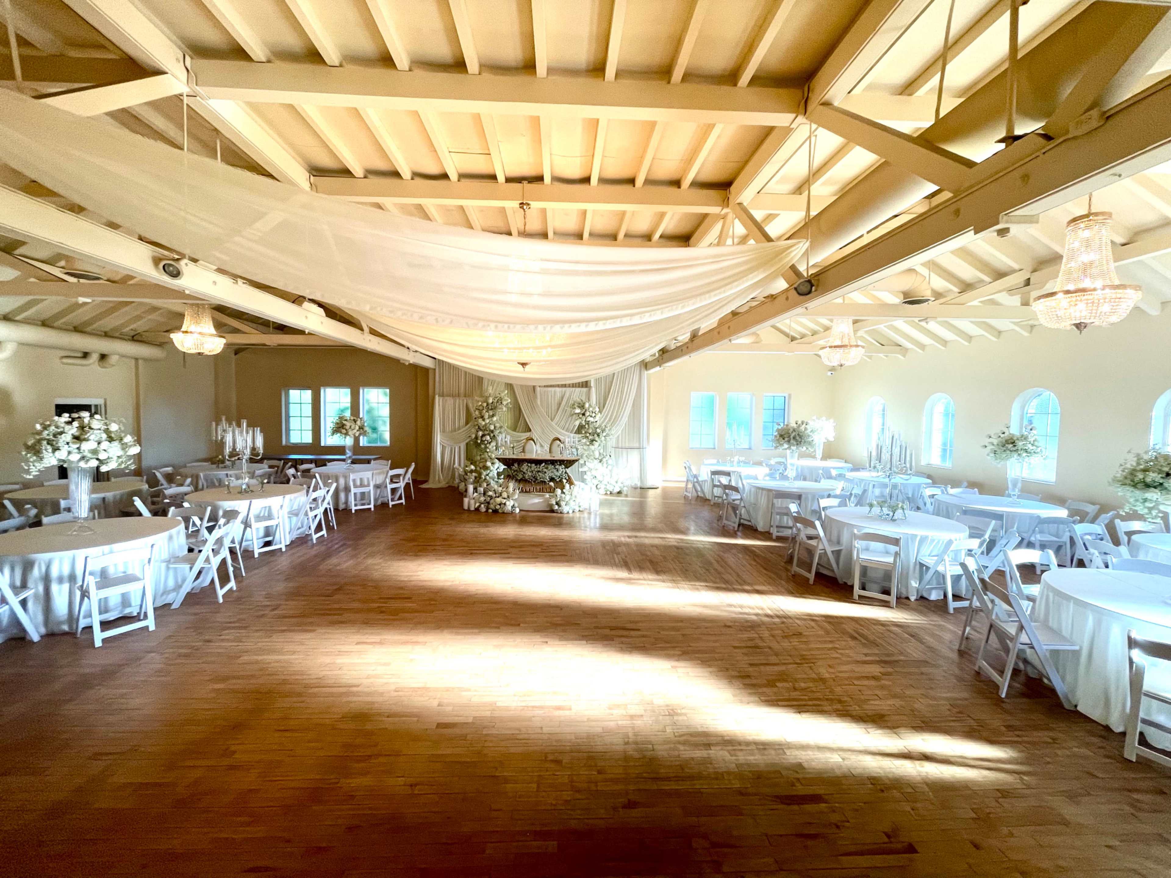 The image shows a spacious, elegant indoor venue with tables set for an event, adorned with white linens and floral centerpieces, under a softly lit canopy.