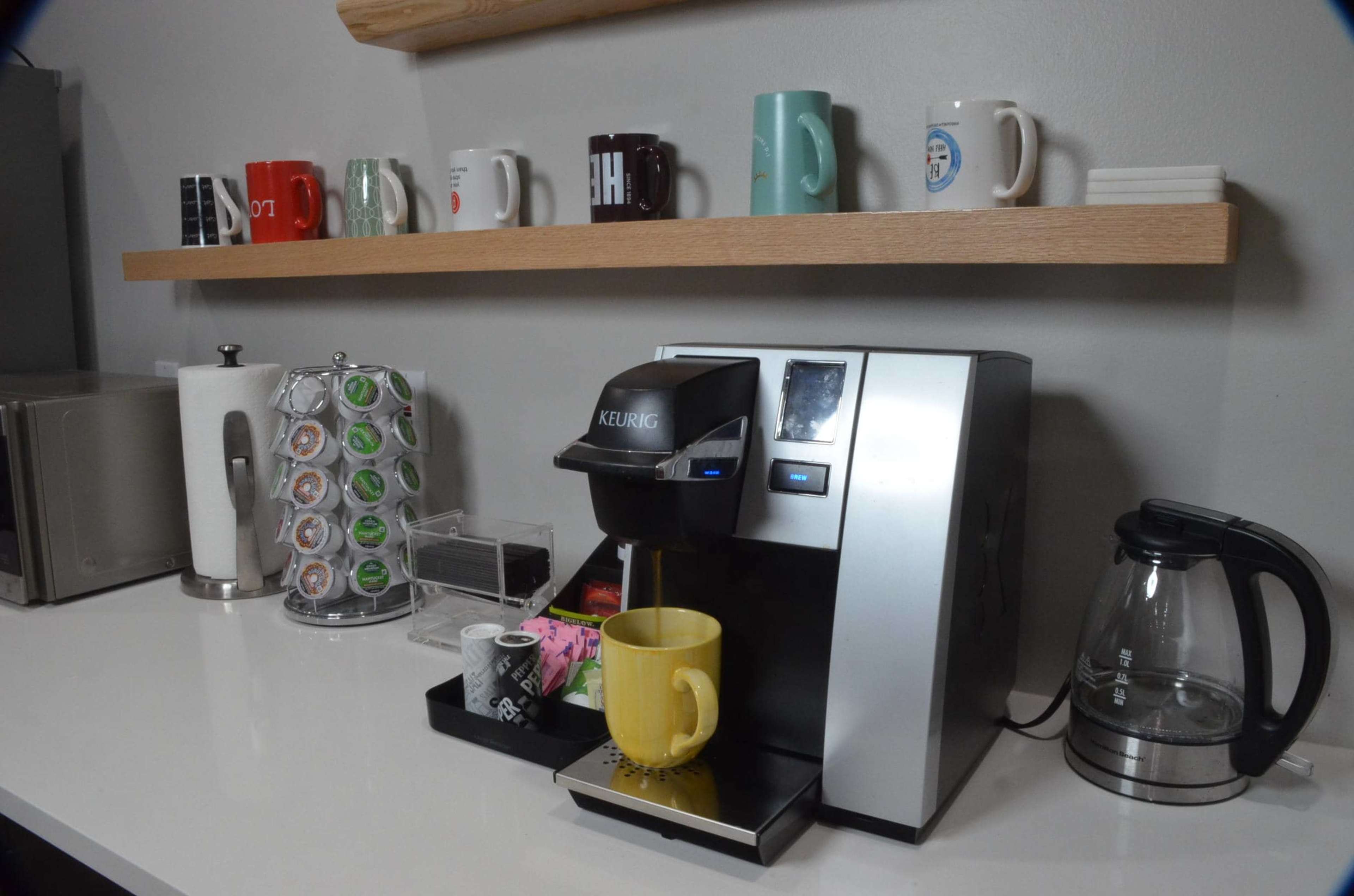 A Keurig coffee maker is positioned on a countertop with a yellow mug under the spout, surrounded by various coffee pods and a row of colorful mugs on a shelf above.