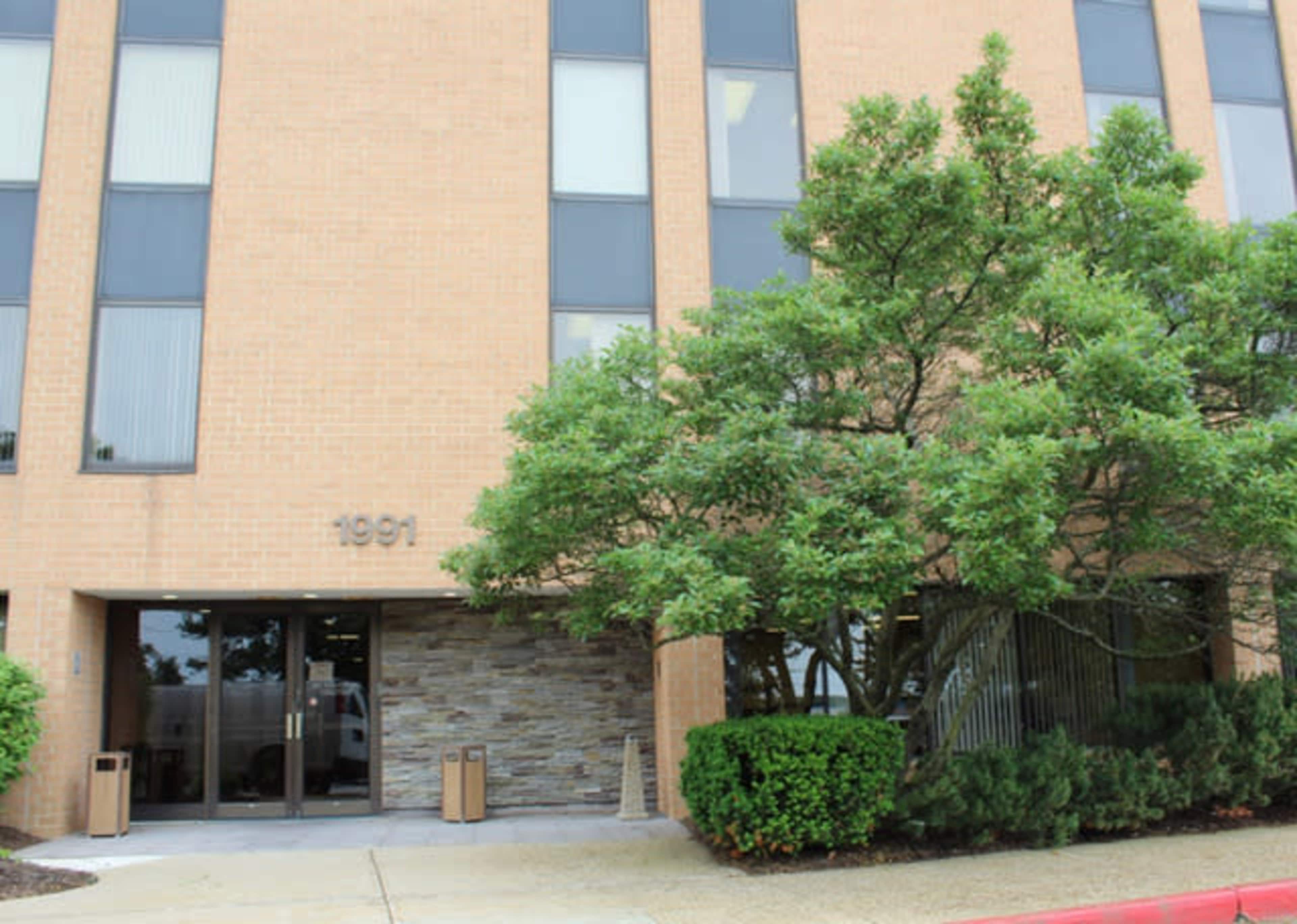 A five-story brick office building with large windows and a landscaped entrance featuring a tree and shrubs.