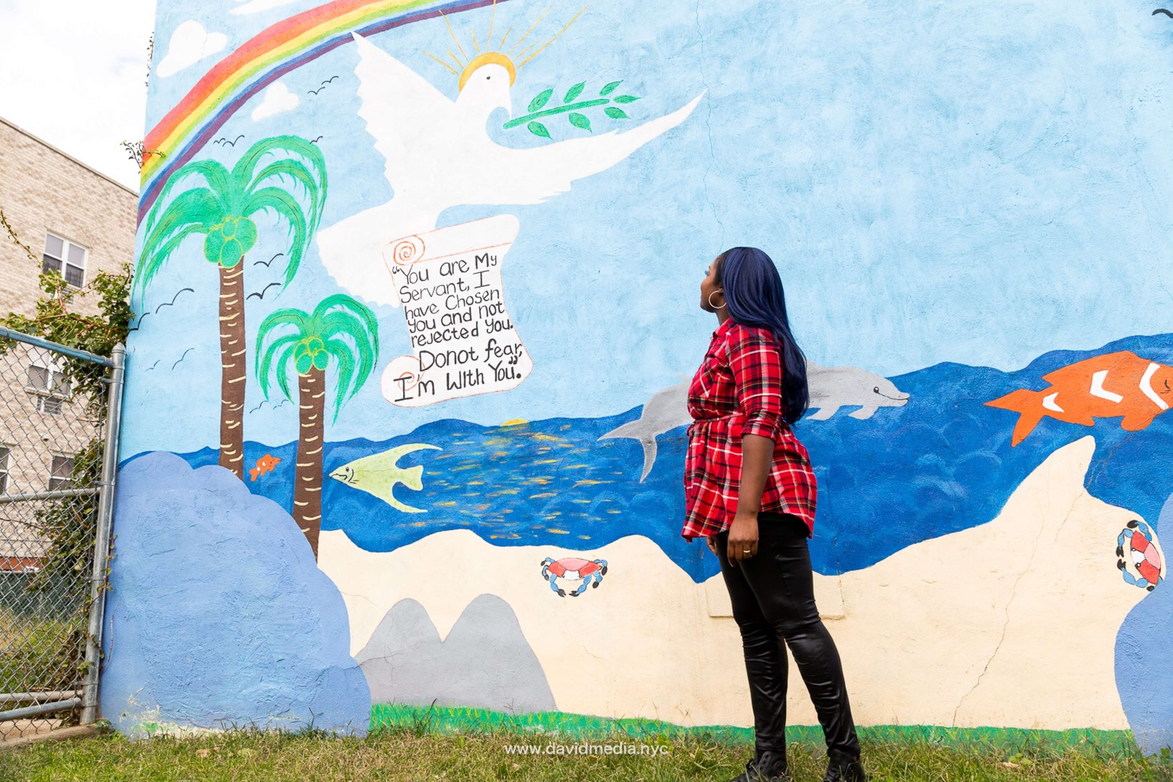 A woman in a checkered shirt stands in front of a colorful mural featuring a rainbow, a dove, and ocean imagery.