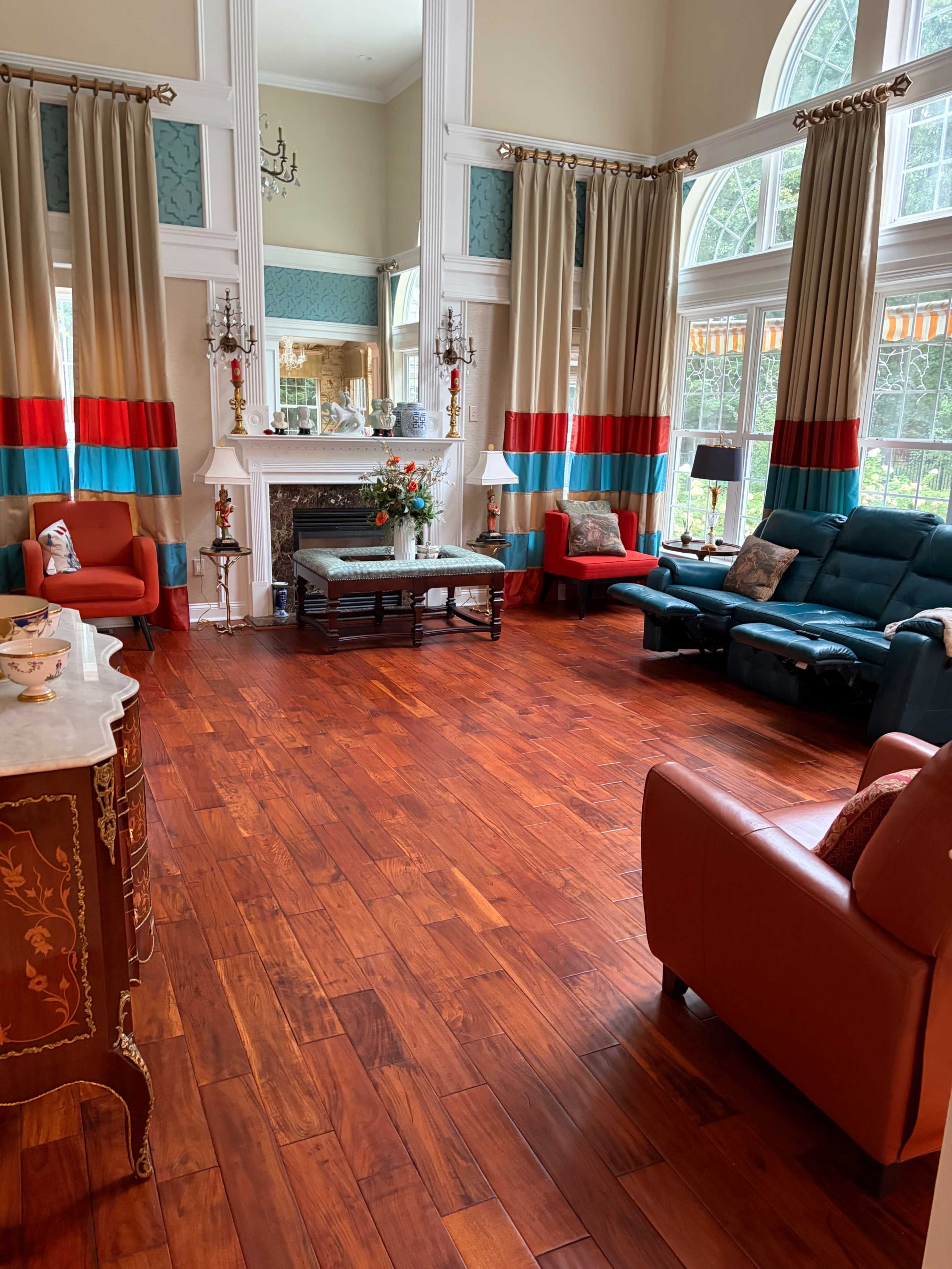 The image shows a spacious living room with large windows, hardwood flooring, and red and blue curtains framing the windows beside various seating arrangements.