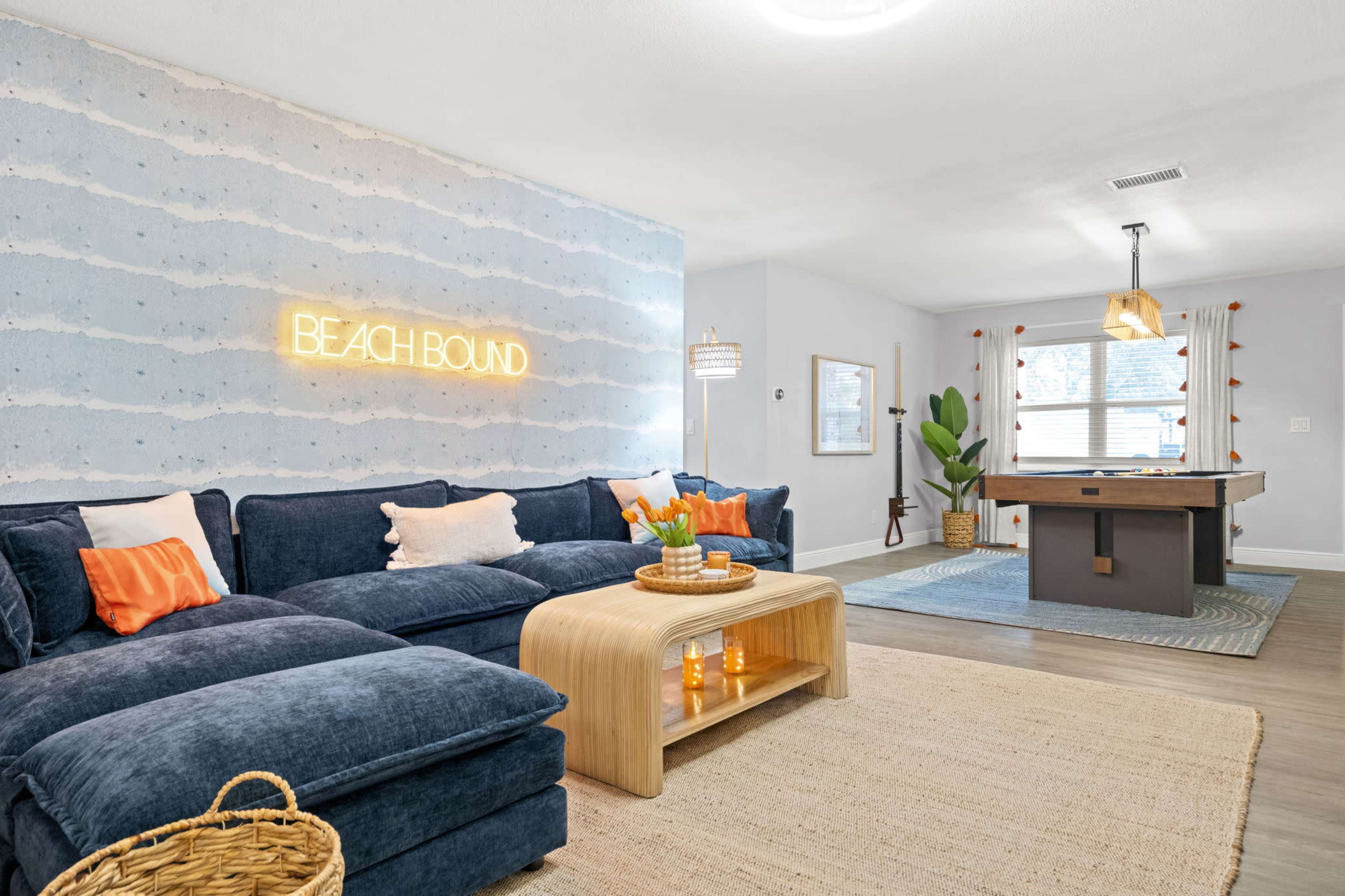 A cozy living room features a navy sectional sofa, a wooden coffee table, and a wall with a "BEACH BOUND" neon sign.