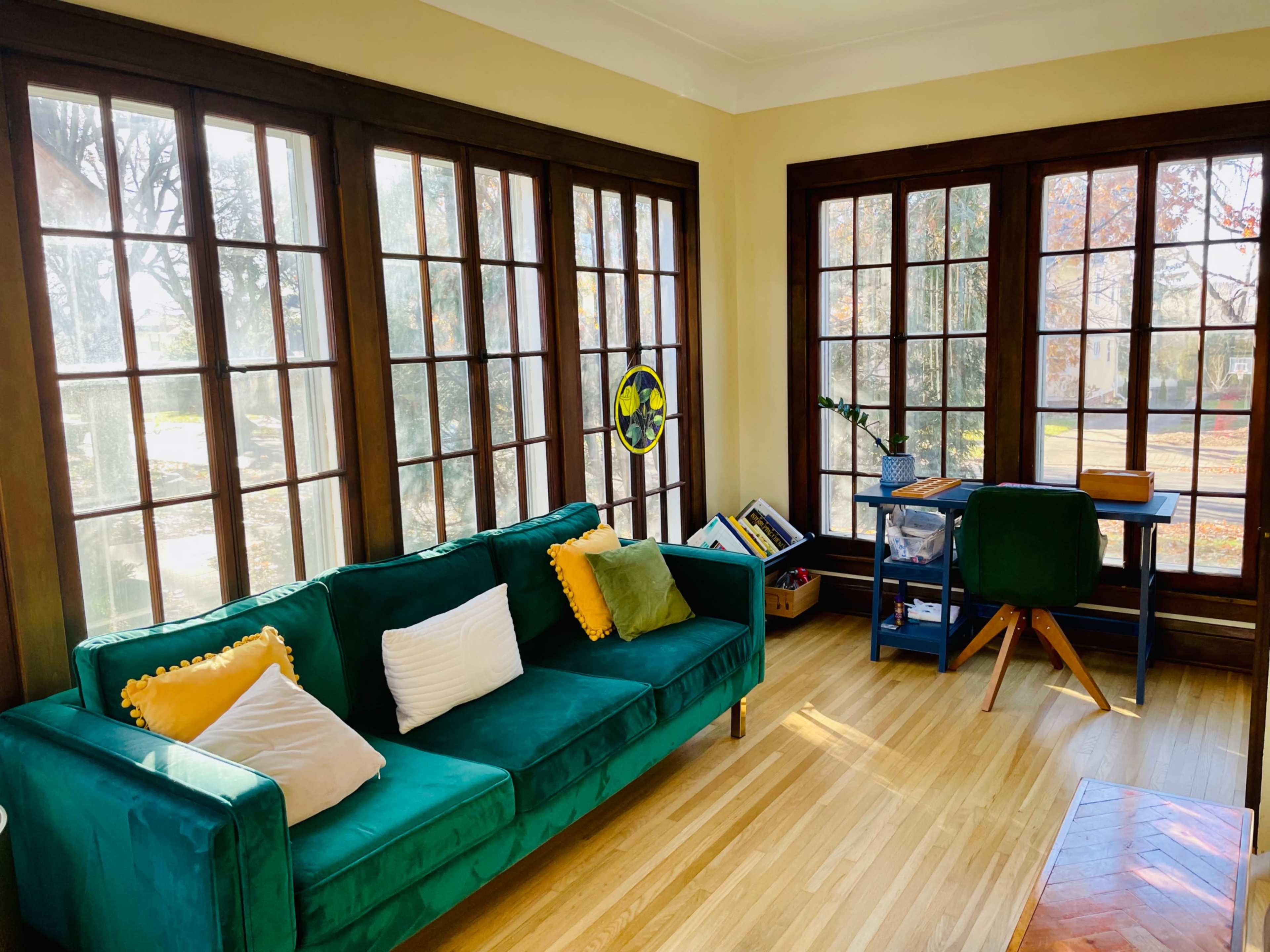 A bright room features a green sofa with colored pillows, a desk, and large windows that allow natural light to fill the space.